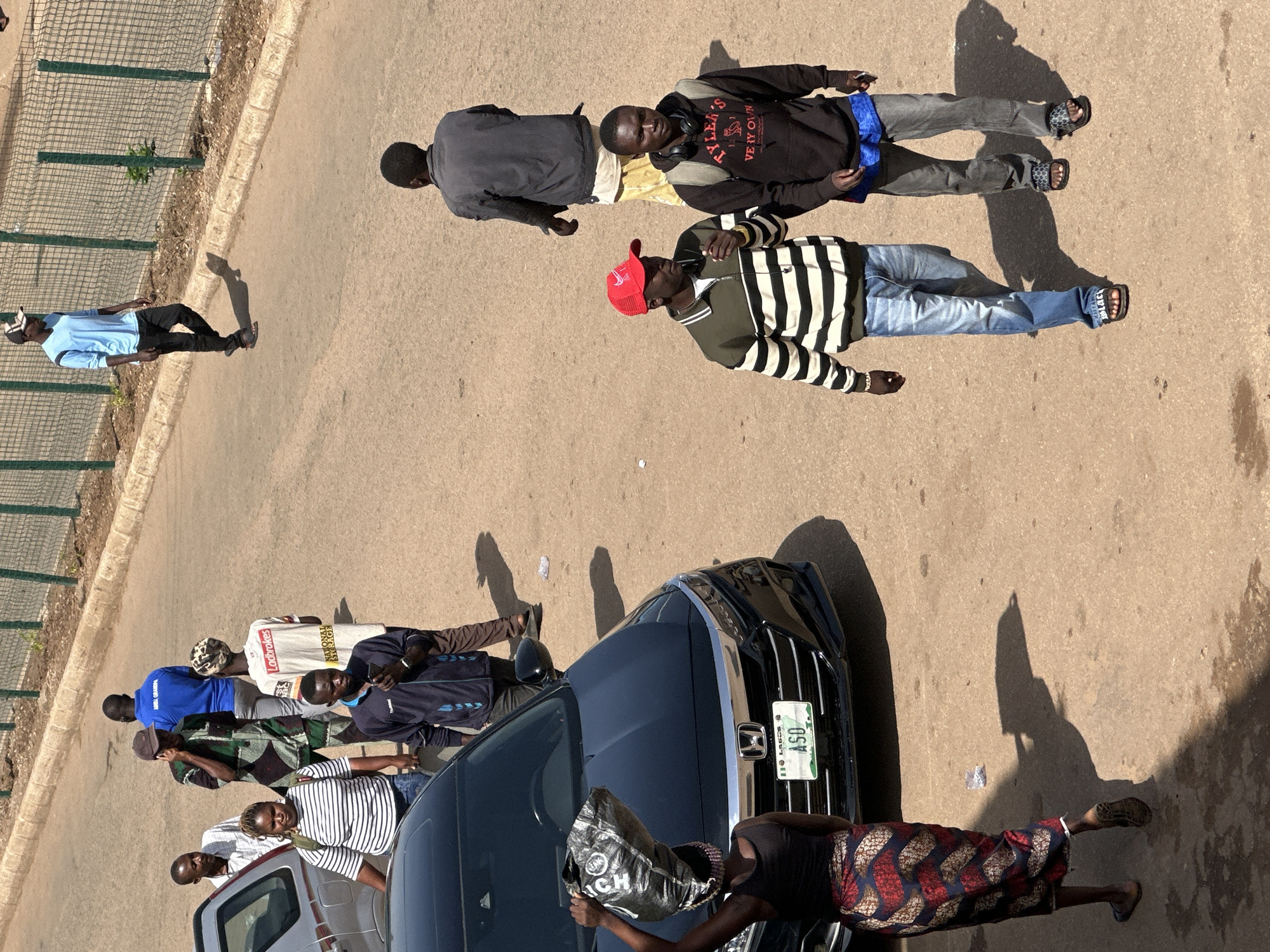 In Bukuru, Nigeria, a street scene unfolds with several individuals. A woman walks carrying a black bag on her head, with a black Honda car parked in front of her. A man in a blue shirt and black pants walks away from the camera near a green metal fence. Another man, also in a blue shirt, is further down the road. Two men are walking towards the camera; one wears a red cap and a striped sweater, while the other is in a black hoodie. Several other individuals are seen walking and standing in the background, some on a paved road and others on a dirt shoulder. The environment appears to be an urban street with buildings and a fence visible.