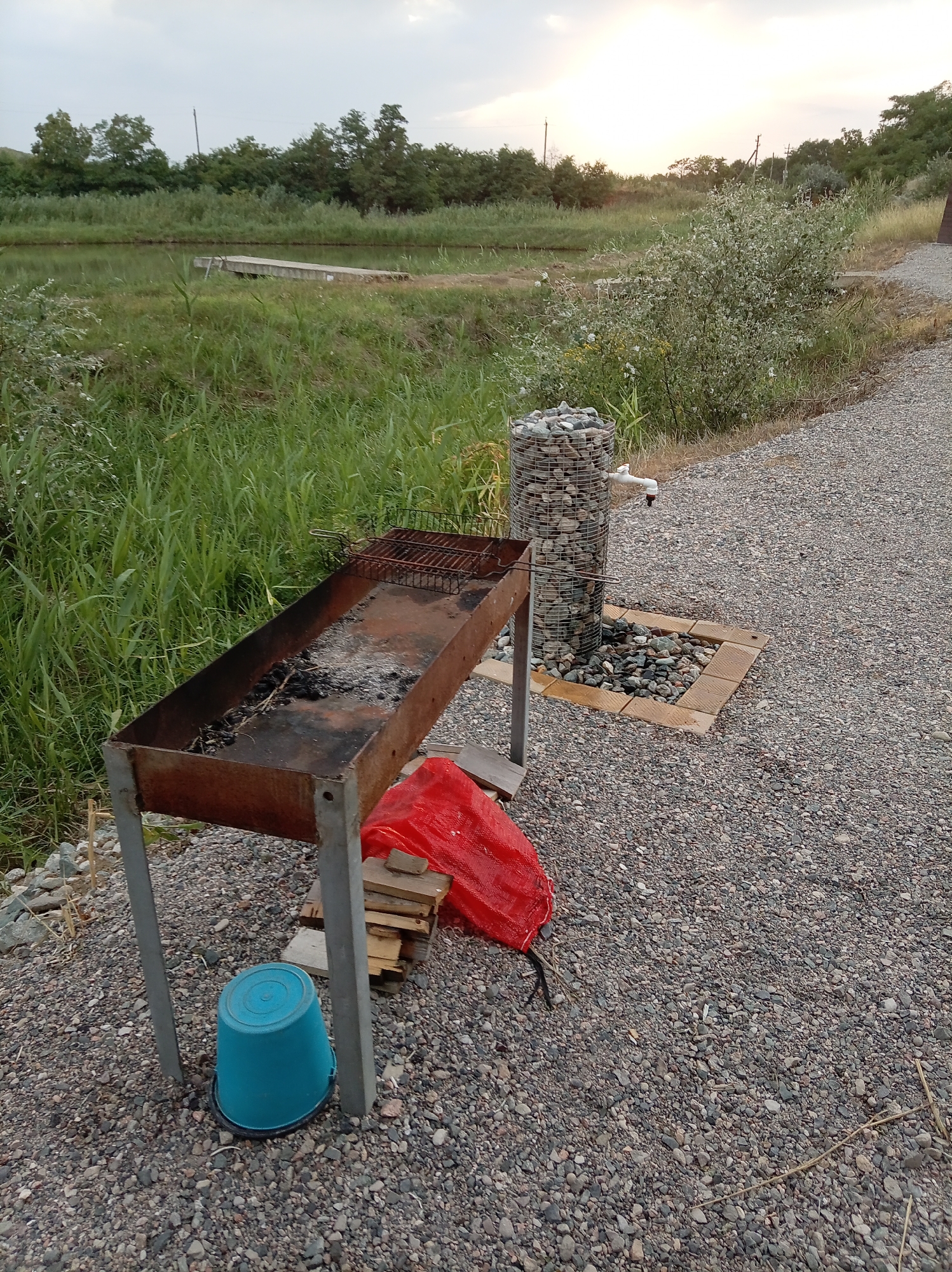 The image depicts an outdoor scene, likely in a rural or semi-rural area. In the foreground, a rusty metal barbecue grill sits on a gravel surface. Beneath the grill, a bright blue plastic bucket is visible, along with a pile of stacked wooden planks and a crumpled red plastic sheet. To the right of the barbecue, a decorative water feature made of a wire mesh cylinder filled with stones stands next to a white faucet. The area around the water feature is bordered by light-colored square tiles.
Behind the barbecue and water feature, a dense area of green grass and reeds stretches towards a body of water. A wooden structure, possibly a small pier or platform, juts out into the water from the bank on the left side of the frame. In the background, a line of trees forms a natural boundary, with the sky above showing a pale, overcast or dusky light. The overall environment suggests an outdoor recreational or picnic area. The location is specified as Гражданское, Russia, which is a settlement within Russia.