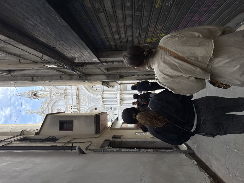 The image depicts a narrow urban alleyway, identified as a *calle* in Venice, Italy, with St. Mark's Basilica visible in the distance at its terminus. Two individuals are walking away from the camera in the foreground. The person on the left wears a black baseball cap, a black jacket, and dark jeans; their brown hair is gathered in a ponytail. The person on the right wears a light-colored trench coat, with brown hair pulled back into a bun, and a brown leather strap from a bag crosses their back.
The alley walls are made of aged masonry. The building on the left features a projecting structure with a small, dark-framed window and an external downspout. The building on the right has a dark, corrugated metal roll-down shutter covering its lower portion, which is defaced with various graffiti tags in multiple colors. Several other indistinct figures are visible further down the alley, preceding the two main subjects. Overhead, a patch of partially cloudy sky is visible between the tall buildings. The ground is a paved surface. The ornate upper facade, spires, and statuary of St. Mark's Basilica are clearly discernible at the end of the *calle*, under the bright sky.
