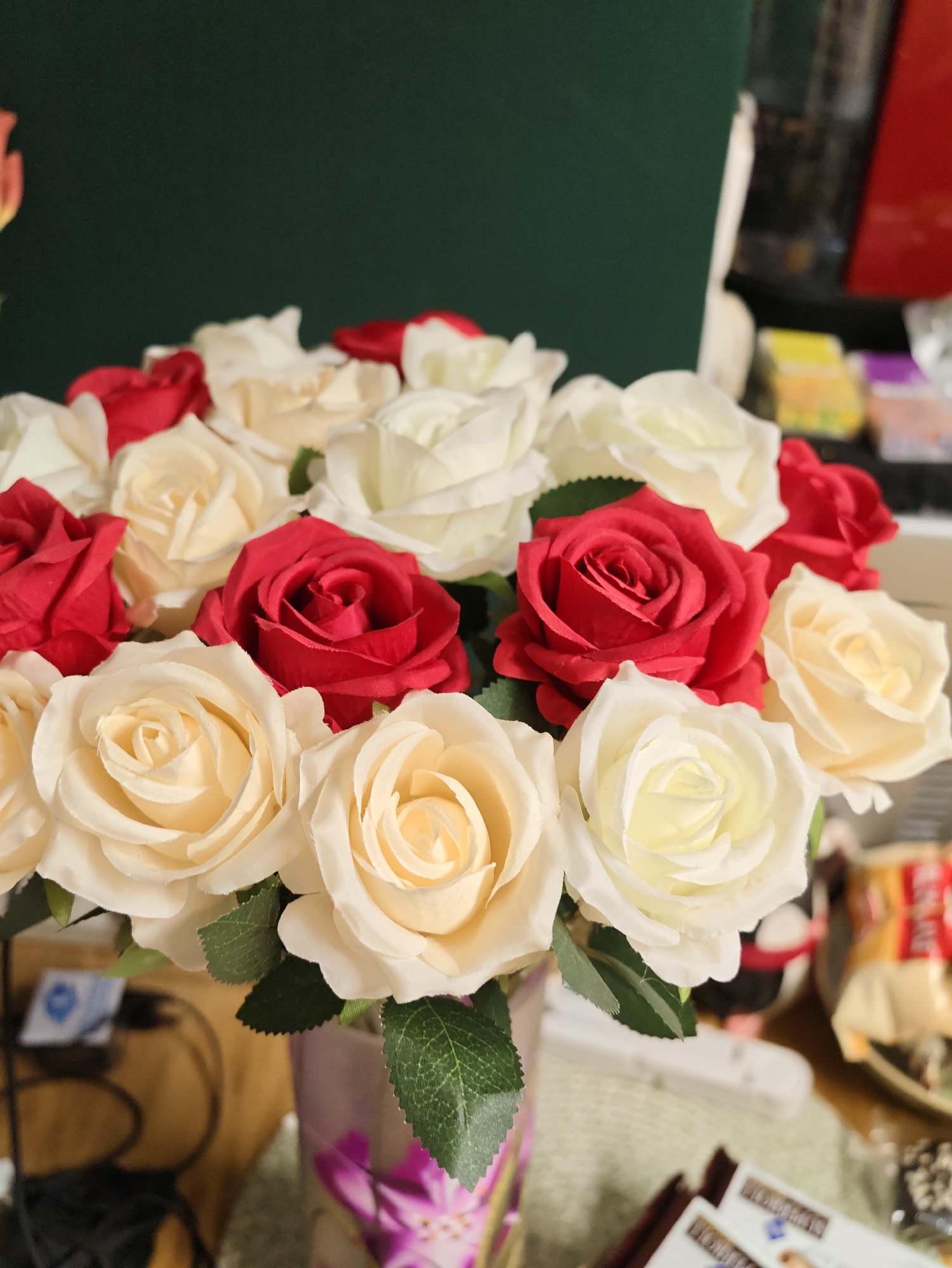 A close-up, eye-level shot captures a bouquet of artificial roses in various shades of red and cream, arranged in a vase with a floral pattern. The flowers are the primary focus, with some appearing more in focus than others. The background is dimly lit and shows a dark green wall on the left, and on the right, shelves with various items, including what appear to be boxes of snacks and possibly toys or figurines. The setting appears to be indoors, possibly a home or a shop, and is not identifiable as being in Petah Tikva, Israel. There are no people visible in the image.