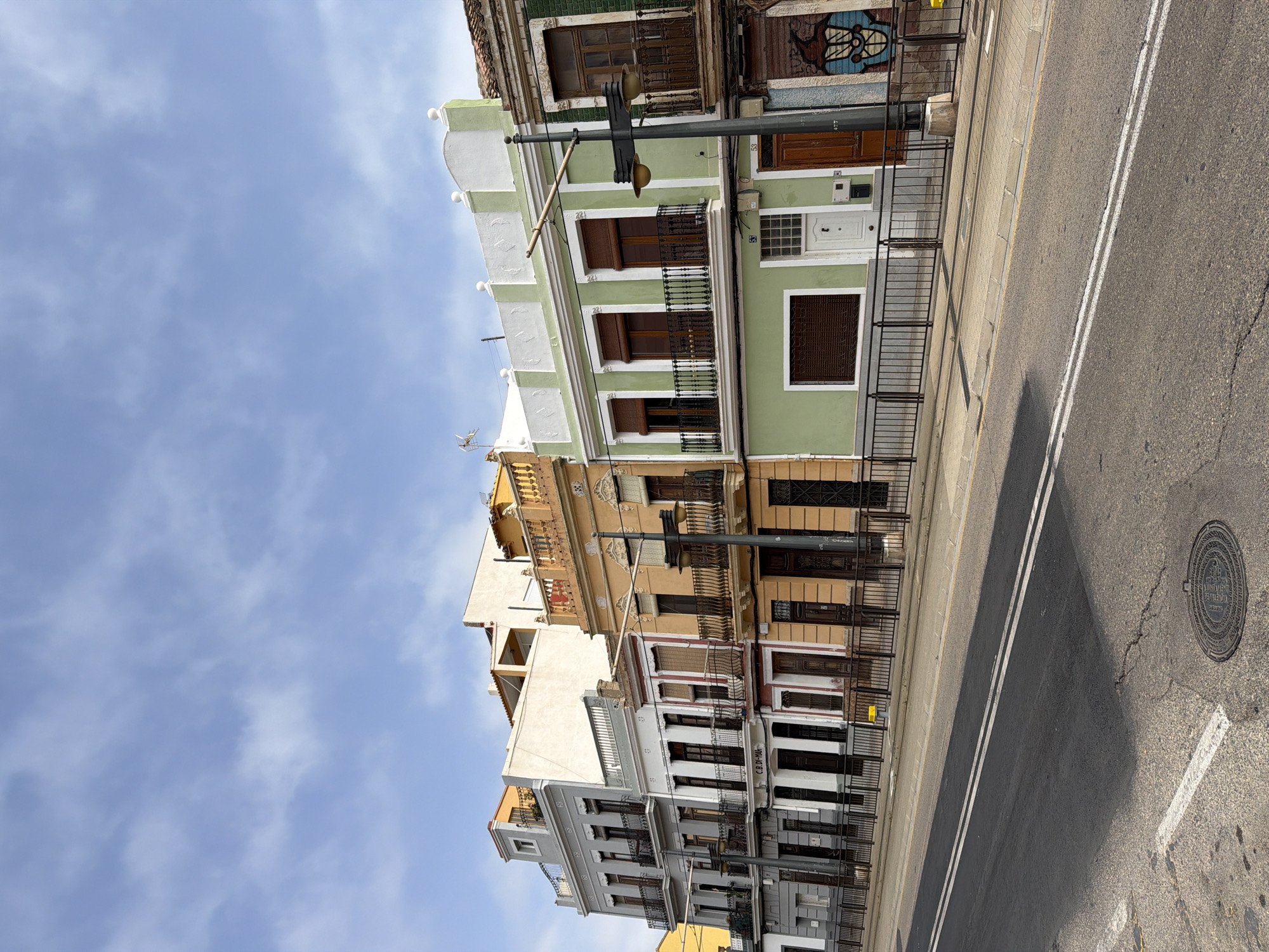The image shows a row of colorful, narrow buildings lining a street in an urban environment. The buildings have multiple stories, with balconies and ornate architectural details. The sky is blue with scattered white clouds. The street in the foreground is asphalt with double white lines and a manhole cover. A black metal fence separates the sidewalk from the buildings. The buildings are painted in various colors, including gray, white, orange, and light green. Some buildings have closed shutters on the windows. There is graffiti on the rightmost building. The overall impression is of a historic street in a Mediterranean city.
