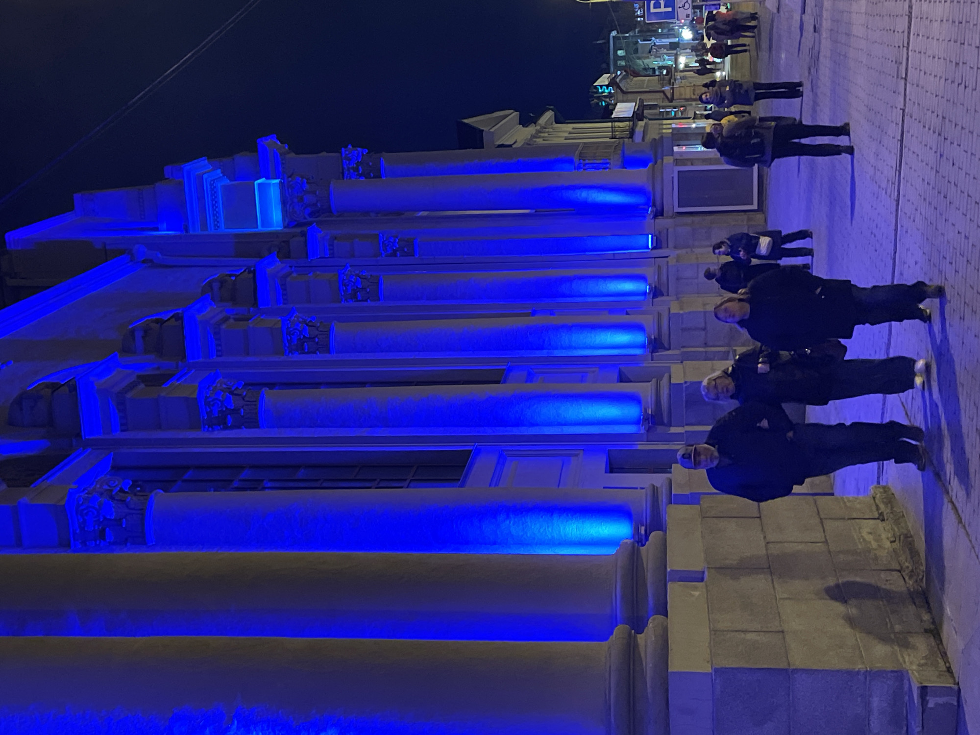 At night, the Ural State University of Economics in Yekaterinburg, Russia, is illuminated with blue lights. The building features a neoclassical design, with large pillars extending the length of the structure. A group of people, dressed in winter attire, are walking along the sidewalk in front of the building. The street is paved with cobblestones. The lighting is bright, but the sky is dark. In the background, street lights and signs are visible, indicating the presence of a commercial area.