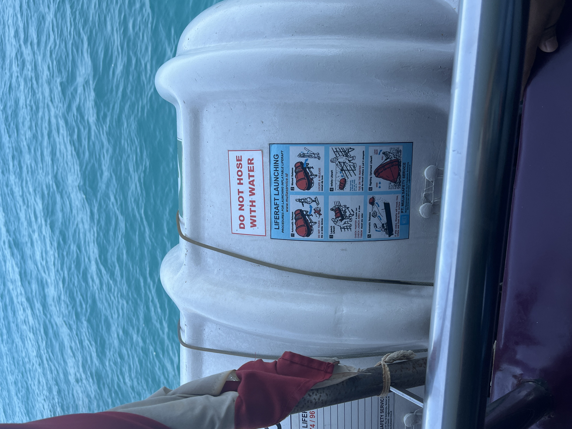 The image shows a liferaft on a boat with the turquoise waters of Karimunjawa, Indonesia, in the background. The liferaft is white and cylindrical with a dark red and white safety instructions label. The label details liferaft launching procedures, including automatic and manual release, inflation, and righting an overturned liferaft. A red and white piece of fabric is draped over a metal pole attached to the liferaft. The bottom of the image shows a metallic railing and a dark-colored boat surface. The waters of Karimunjawa are calm and appear to be shallow.