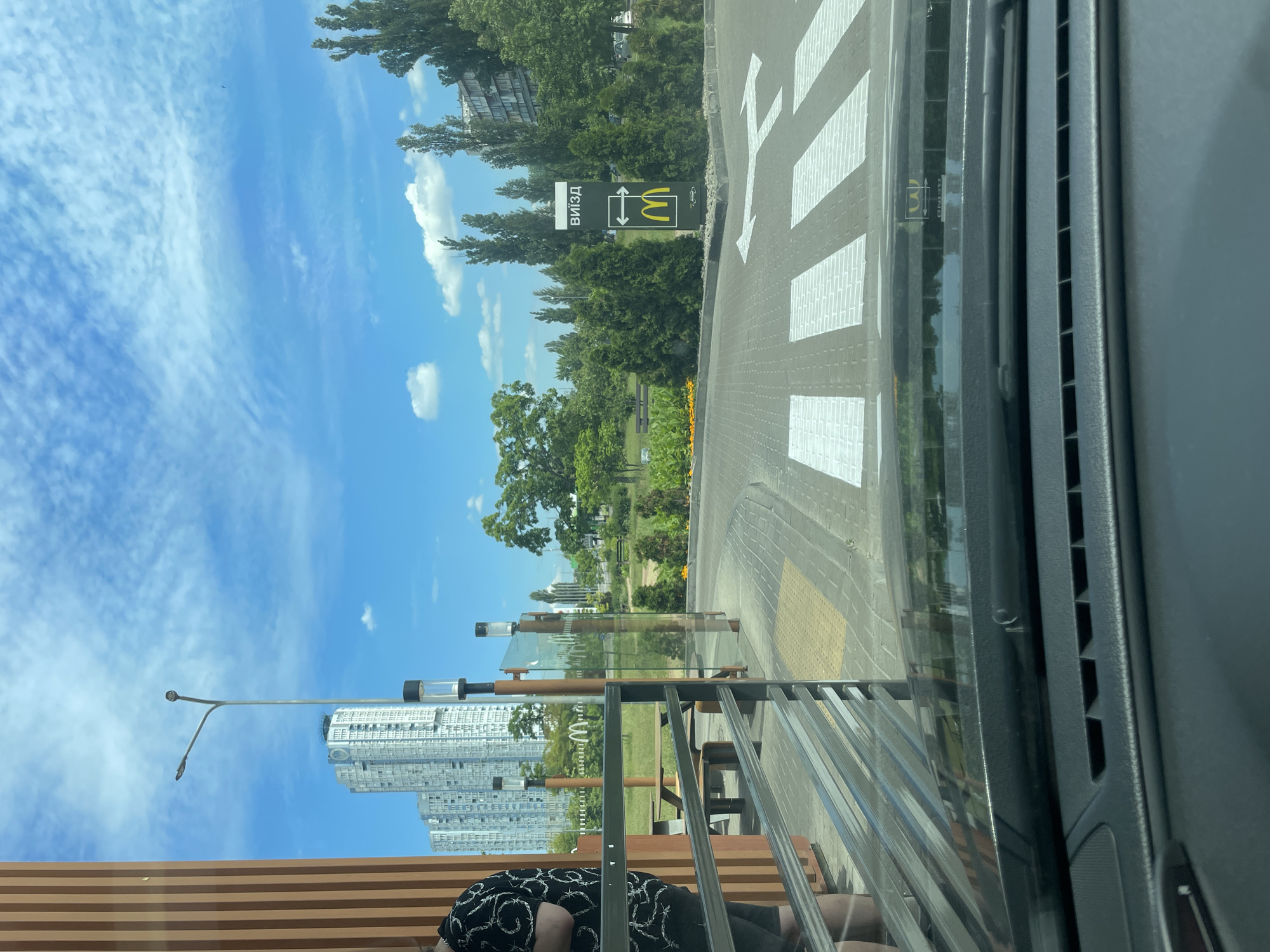 The image shows an outdoor scene viewed from inside a vehicle. The camera is angled to the side. In the foreground, a person's legs and lower torso are visible, wearing dark shorts with a white pattern. They are sitting on a wooden bench with horizontal slats, and a metal railing is in front of the bench.
Beyond the bench, there is a paved area with white crosswalk markings, indicating a pedestrian crossing. A street sign with the McDonald's logo and text in Cyrillic that reads "ВИЗД" (meaning "entrance") is visible to the left of the crosswalk. Trees and greenery line the area, with a modern, tall building in the background.
The sky is blue with scattered white clouds, suggesting it is daytime with fair weather. The overall setting appears to be urban, possibly near a McDonald's restaurant in Kyiv, Ukraine, given the text on the sign. The scene captures a moment of rest or waiting, perhaps before or after a meal. The reflection in the car window shows a slightly distorted view of the surroundings.