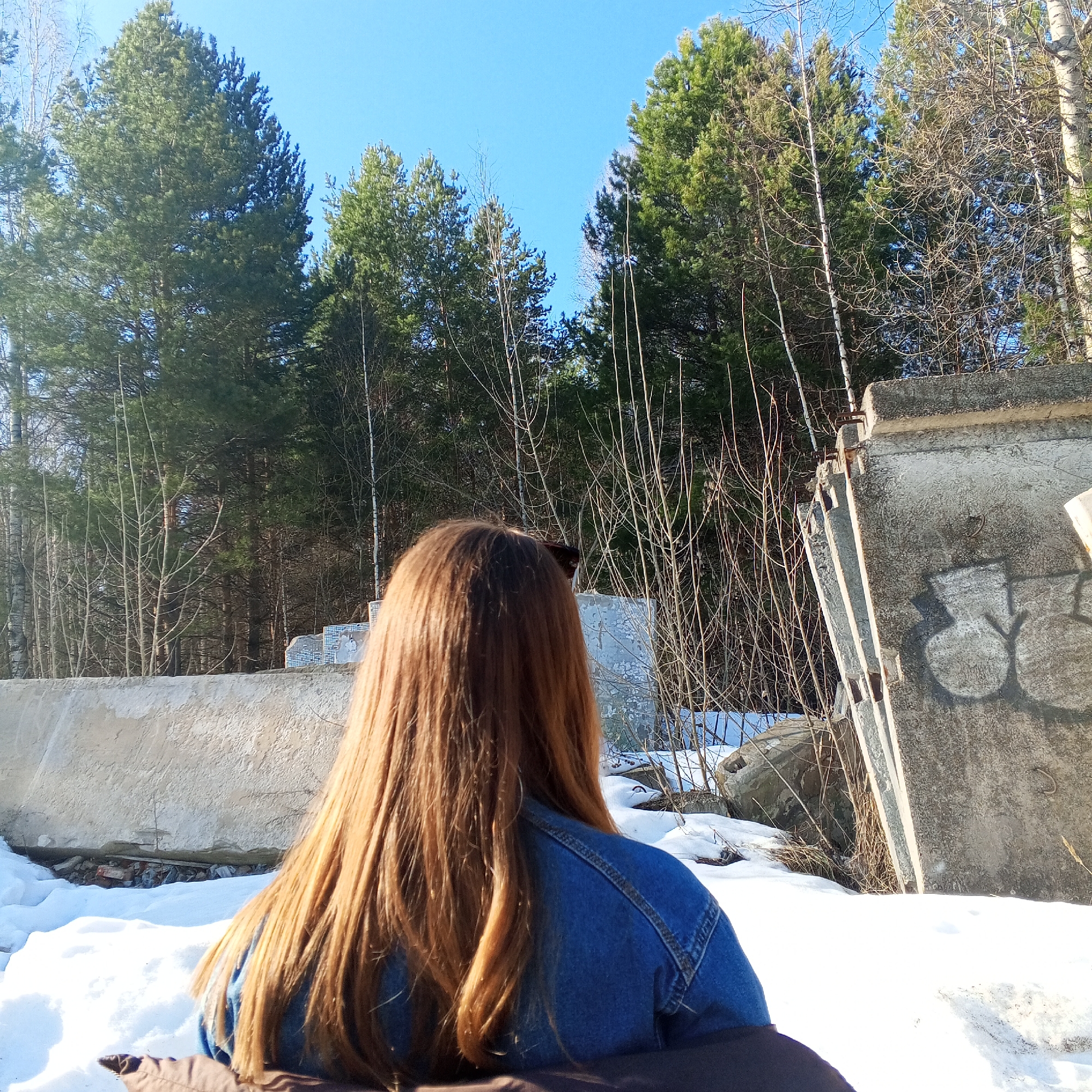 A woman with long, reddish-brown hair, seen from the back, is wearing a blue denim jacket. She is in a snowy area next to what appears to be a partially demolished concrete structure or set of concrete barriers with graffiti. The background shows a dense forest of evergreen trees and bare-branched trees under a clear blue sky. The location is likely the city of Chaikovsky, Russia, but specific location details are not provided.