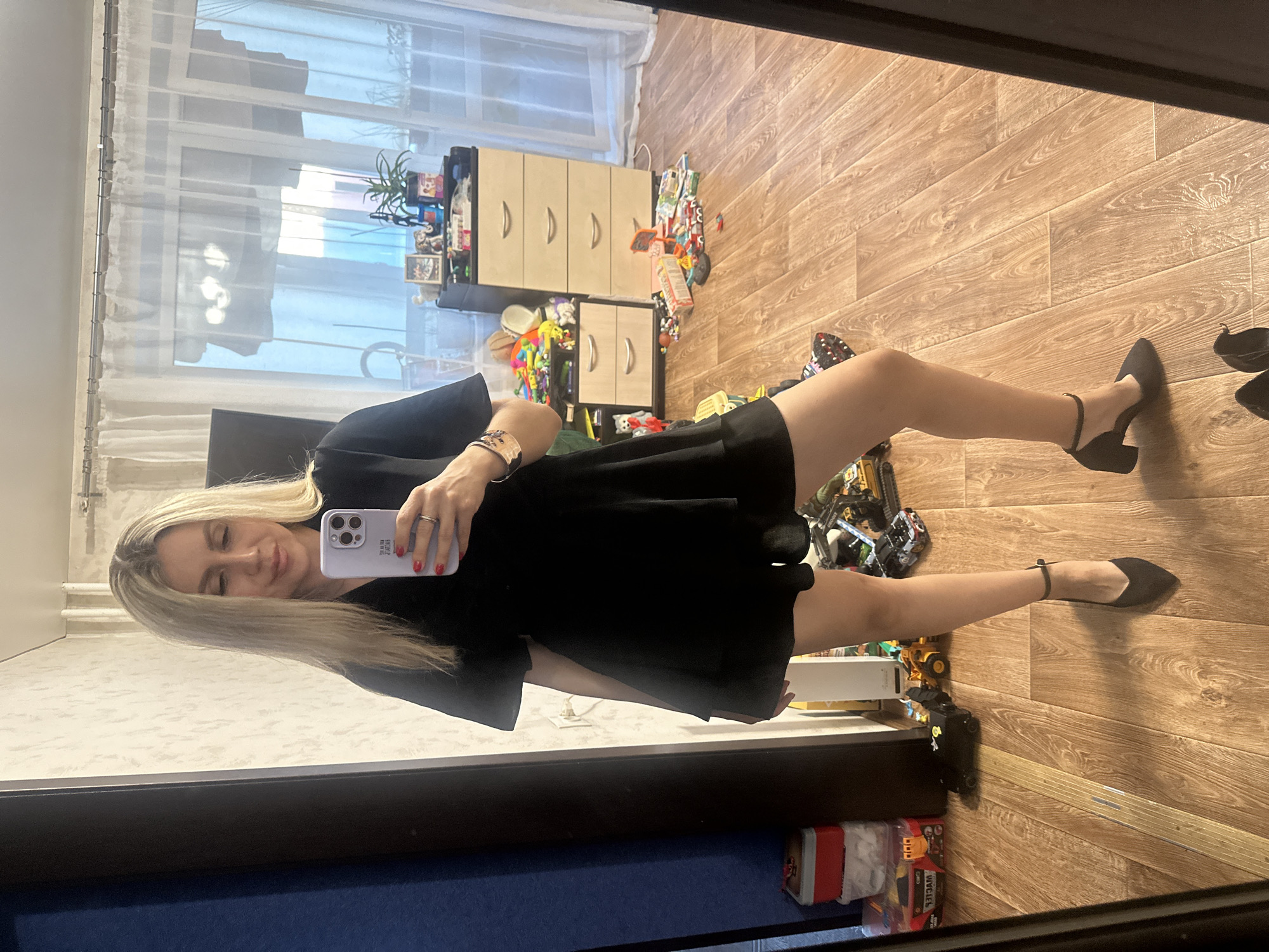A woman stands in a room, taking a mirror selfie. She has blonde hair and is wearing a black dress and black heels. The room appears to be a bedroom or living area, with wooden floors, a dresser, and various toys scattered on the floor. Curtains are visible in the background. The overall scene suggests a casual, domestic environment.