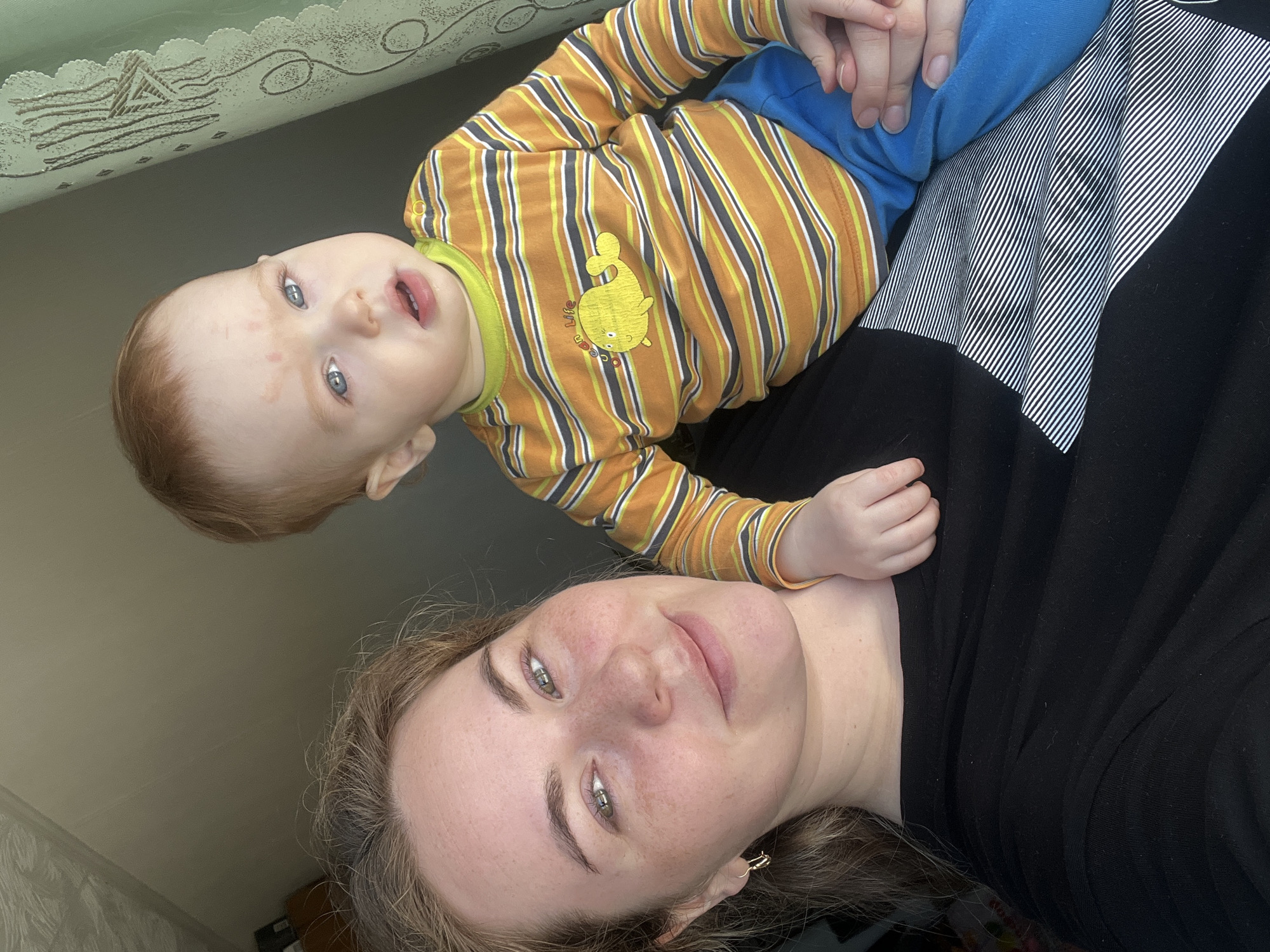 A woman and child are shown in a close-up portrait. The woman lies on her back, wearing a black shirt. A young child, facing forward, is positioned on the woman's chest; the child wears a yellow-and-brown striped shirt with a yellow whale on the chest, as well as blue pants. The child's hand rests on her chest. The location appears to be indoors in a house in Сосново, Russia, near a light wall and a surface covered with a printed fabric.
