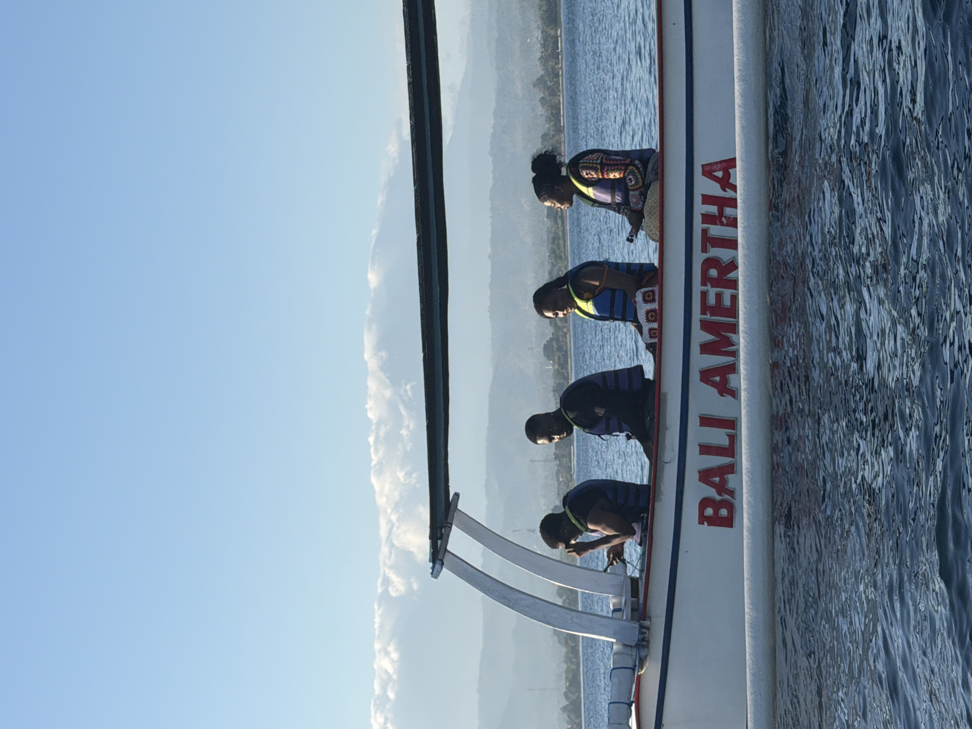 An outdoor, low-angle, medium shot captures four people in life vests on a white boat with a blue stripe and red lettering that reads "BALI AMERTHA." The boat is on the water, with distant mountains and a bright blue sky with scattered clouds in the background. The person on the far left is a Black man wearing a black t-shirt and a blue life vest, looking at his phone. Next to him is another Black man in a black t-shirt and blue life vest, looking directly at the camera. The third person is a Black woman with dark hair, wearing a black top and a colorful life vest with a patterned design on the sleeves and shoulders, looking to her left. The fourth person, a Black woman with her hair pulled back, is wearing a black top and a life vest with a colorful pattern on the chest, looking to her right. The scene is likely in Seririt, Indonesia, given the boat's name.