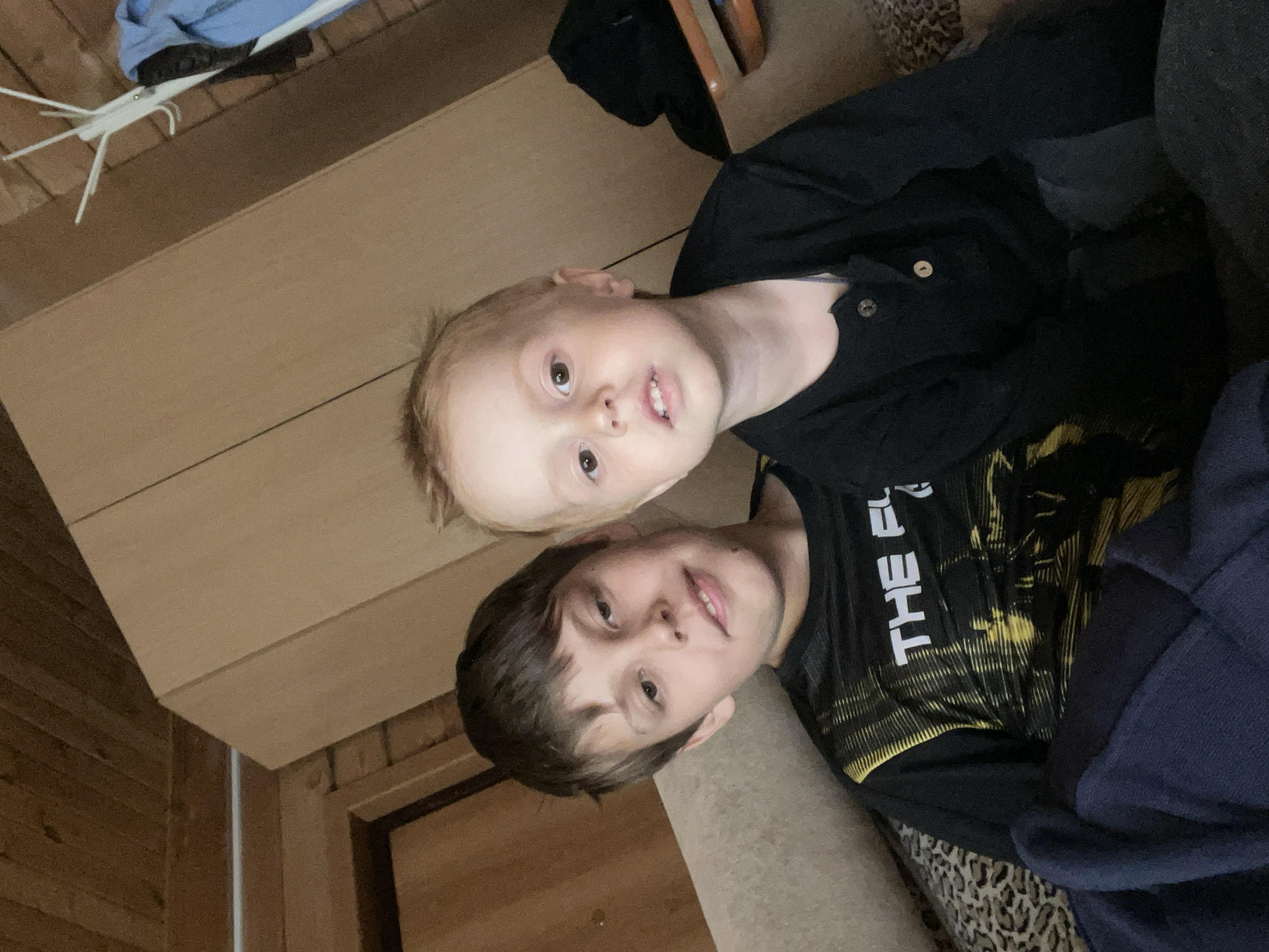 An indoor portrait taken in Chaikovsky, Russia, features two boys, likely siblings, looking towards the camera. The image is oriented vertically, with the subjects appearing to be lying down or leaning back. The younger boy, with light brown or blond hair, is positioned above the older boy. He wears a dark, possibly navy, collared shirt with buttons and has a neutral expression with his mouth slightly ajar. The older boy, with darker hair, is wearing a black t-shirt with a striking yellow, possibly abstract or graphic, pattern. White text reading "THE F" is visible on his shirt, suggesting part of a larger word or phrase. He has a calm expression with a faint smile.
The setting appears to be a cozy home environment. In the background, to the upper left, there is a wooden wall paneling and a light-colored wooden surface, possibly a shelf or part of a piece of furniture. A white clothes rack or drying rack with some clothing items is also visible in this area. To the right of the older boy, a dark blue fabric, possibly a blanket or another piece of clothing, is present, resting on a surface with a visible leopard print pattern. The overall lighting suggests an indoor shot, possibly under natural light, and the expressions of the boys are relaxed.
