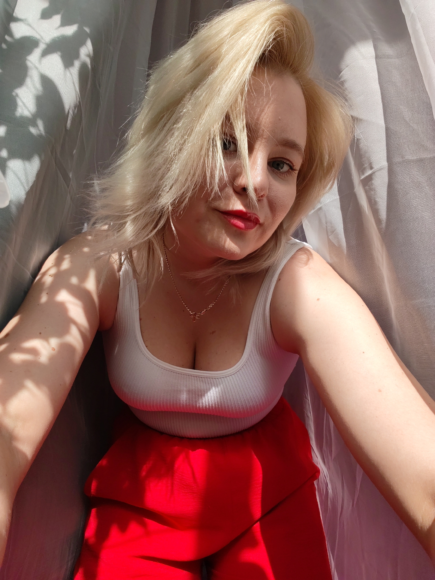 A light-skinned woman with blonde hair is pictured from a high-angle perspective in an indoor environment. She is wearing a white tank top and a red skirt or pants. She is also wearing red lipstick and a delicate necklace. The background is draped with what appears to be a translucent white or light grey fabric, and shadows cast by foliage or a plant are visible on the fabric to the left of the frame. There is no identifiable location of Izhevsk, Russia, in the image.