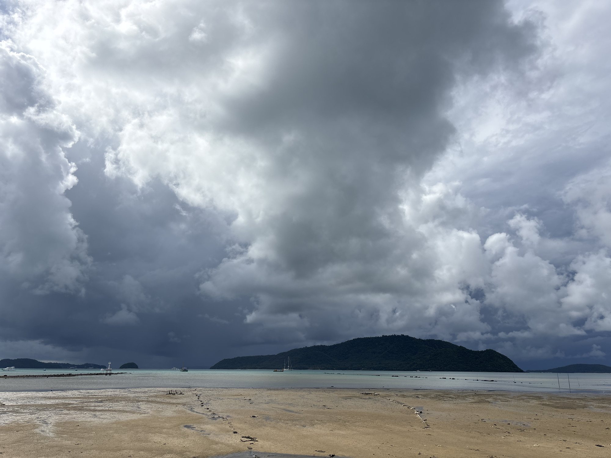 The image depicts a beach scene in Rawai, Thailand, under a dramatic, cloudy sky. The foreground is a sandy beach with tidal pools and footprints visible. In the middle ground, the calm, shallow water stretches towards a distant, tree-covered island. Several boats are anchored in the water, and a lighthouse or marker stands near the shore. The background is dominated by a dark, brooding cloud formation, suggesting an impending storm or the aftermath of one. The overall atmosphere is one of natural beauty tinged with a sense of foreboding due to the weather.