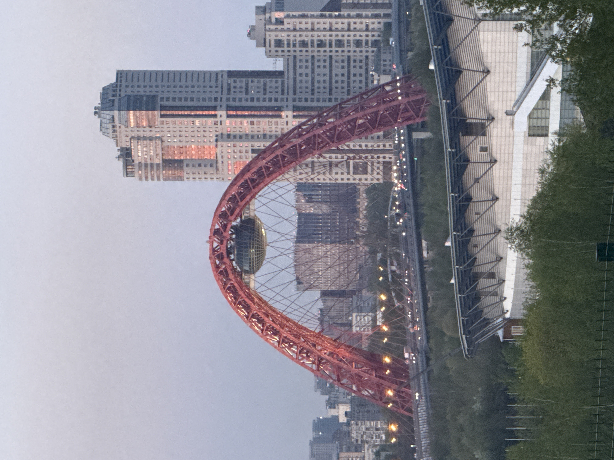 The image depicts a red, arch-shaped bridge spanning a river in an urban setting. The bridge features a spherical structure at the apex of the arch. High-rise buildings are visible in the background, suggesting a densely populated area. A major road with vehicular traffic runs alongside the river. Dense greenery is present along the riverbank, providing a contrast to the concrete and steel of the city. The location is identified as Khimki, Russia, based on the distinctive architecture of the bridge and surrounding buildings. No people are visible in the image.