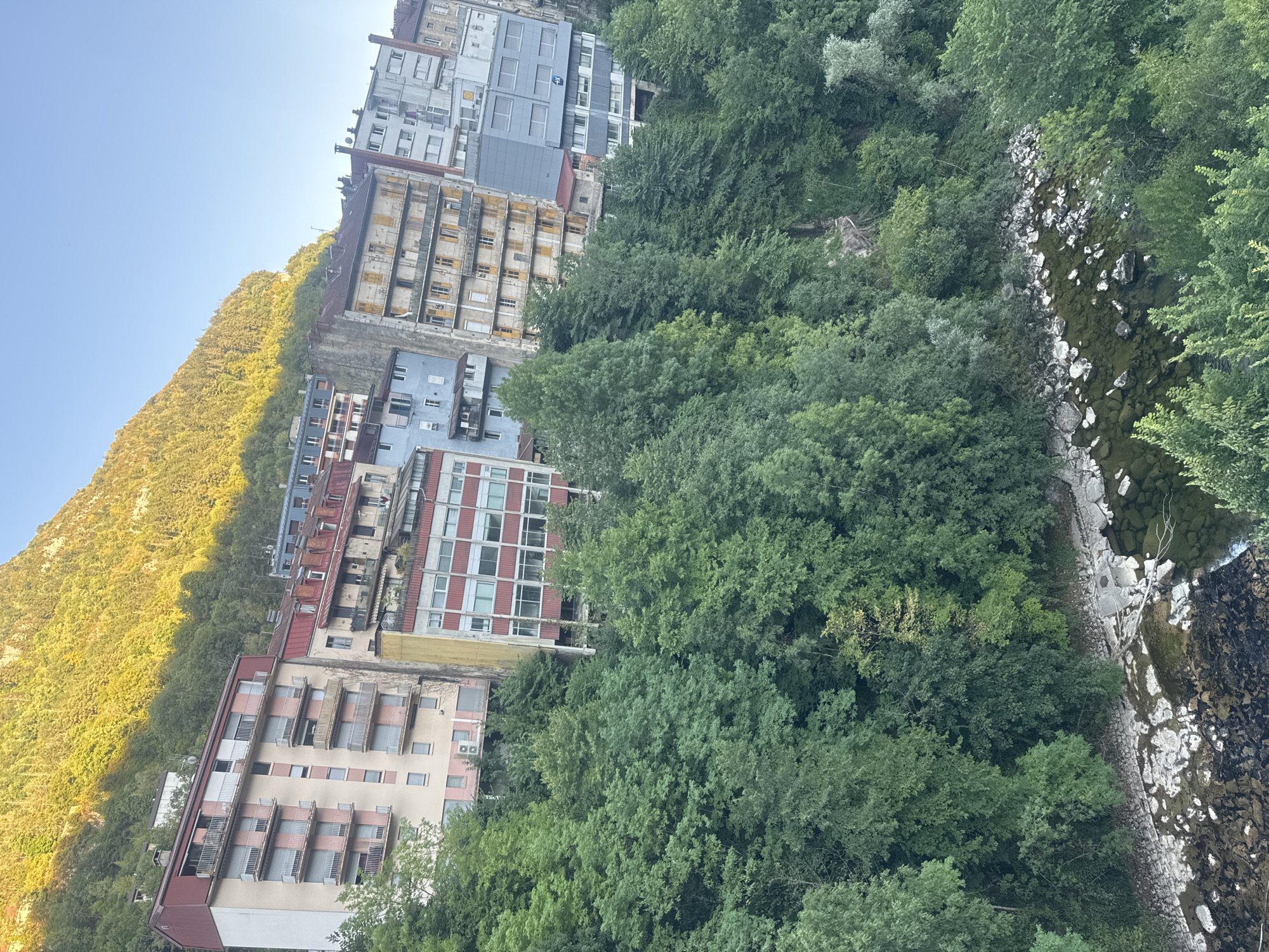 In Saint-Claude, France, multi-story apartment buildings are nestled into a steep hillside, overlooking a rocky river. The architecture varies, with some buildings featuring a pastel pink and white facade, while others display a more muted gray or red exterior. Balconies with railings are visible on many of the buildings, and some have closed shutters. Lush green trees and foliage cover the lower slopes of the hill, contrasting with the urban structures. The river below is shallow and clear, with visible rocks and the dark, rippling water indicating flow. The sky is a clear blue, and the sun casts a warm glow on the upper portions of the wooded hillside.