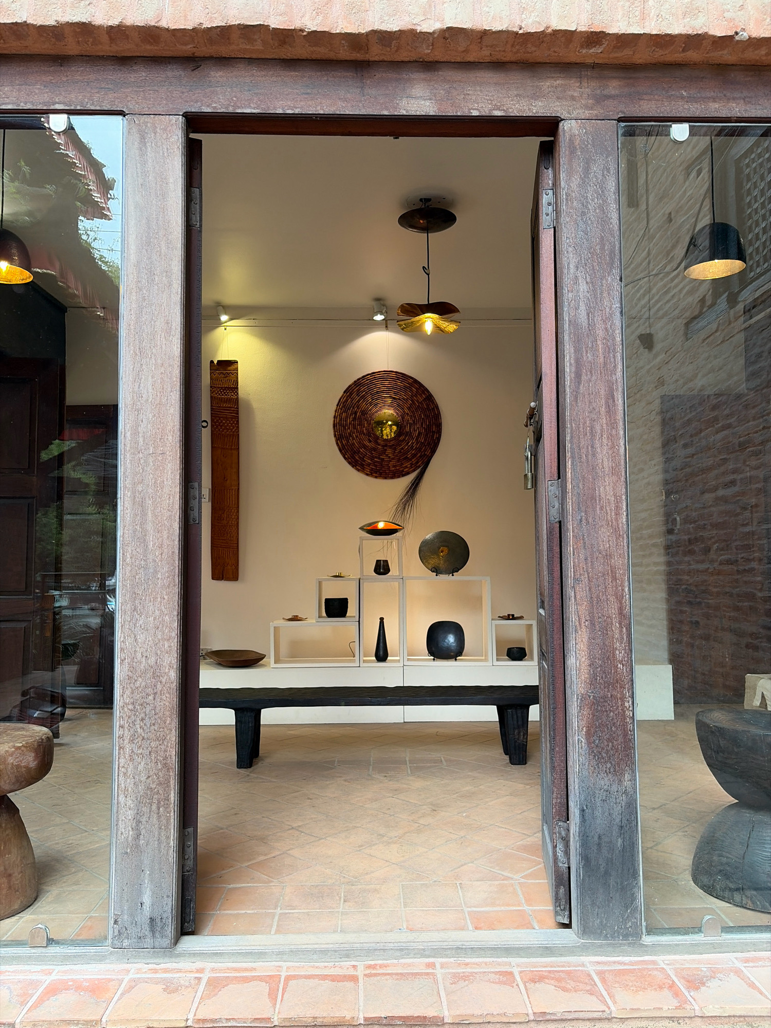 The image shows the entrance to an indoor space, possibly a shop or gallery, in Lalitpur, Nepal. The entrance is framed by weathered wooden doors and glass panels. Inside, the space is brightly lit with artificial lights. Various handcrafted decorative items are displayed on shelves and walls, including woven art pieces, ceramic vases, and wooden sculptures. A low black bench is placed in front of a set of white shelves, which are arranged in a stepped pattern. The floor is tiled with terracotta-colored squares. To the left of the entrance, a wooden stool is partially visible. The overall impression is of an art or craft store showcasing traditional Nepalese artistry.