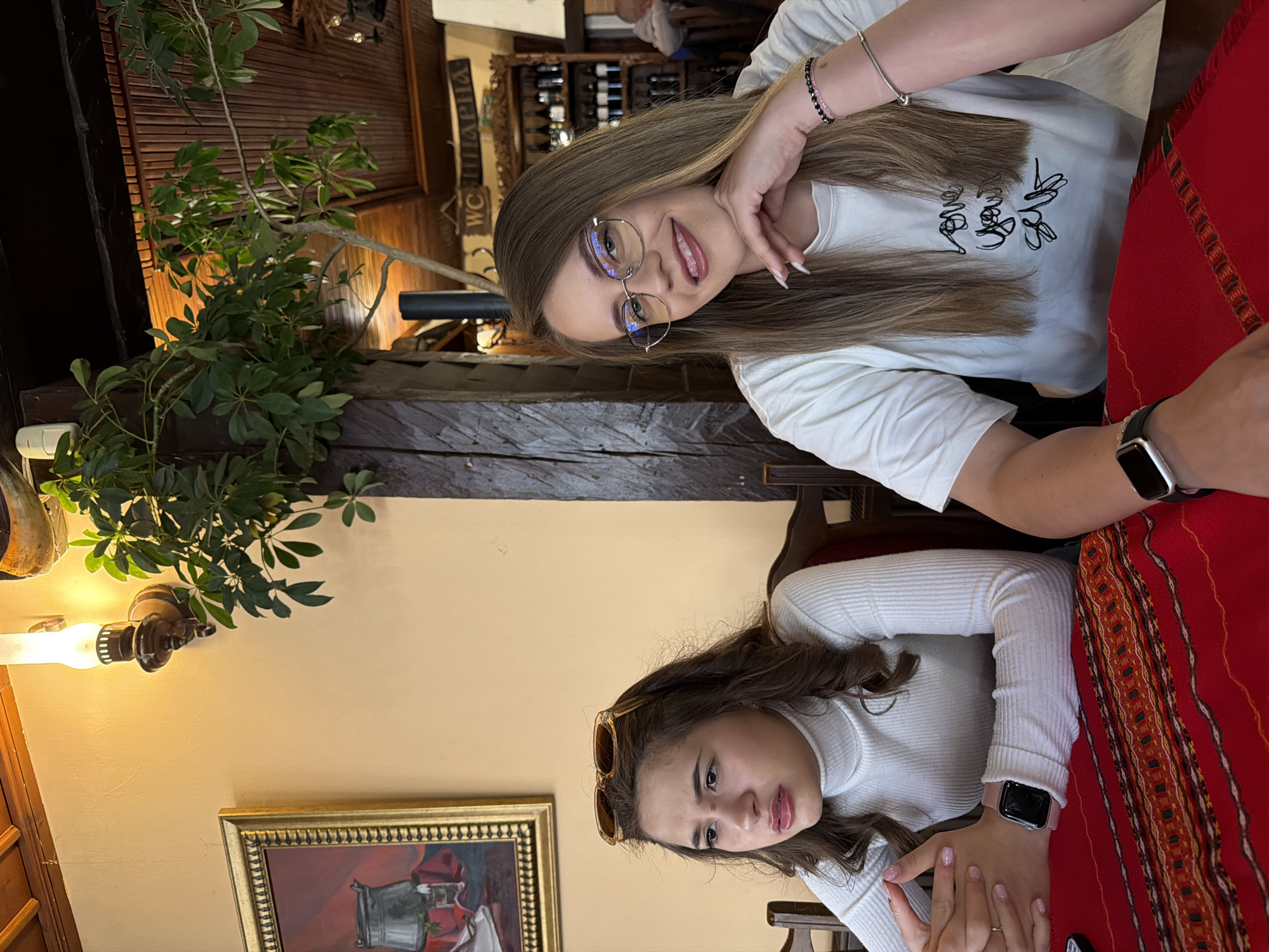 Two women are seated at a table indoors. The woman at the top of the image has long brown hair, glasses, and is wearing a white t-shirt. The woman at the bottom of the image has short brown hair and is wearing a white turtleneck. They both appear to be posing for the photograph. The table is covered with a red patterned tablecloth. The location is a restaurant in Panagyurishte, Bulgaria, suggested by the style of the interior decor, including a dark wooden beam, an ornate picture frame, and possibly the style of the red tablecloth. The interior features wooden furniture and decorative elements such as a lamp with foliage and wine racks in the background.