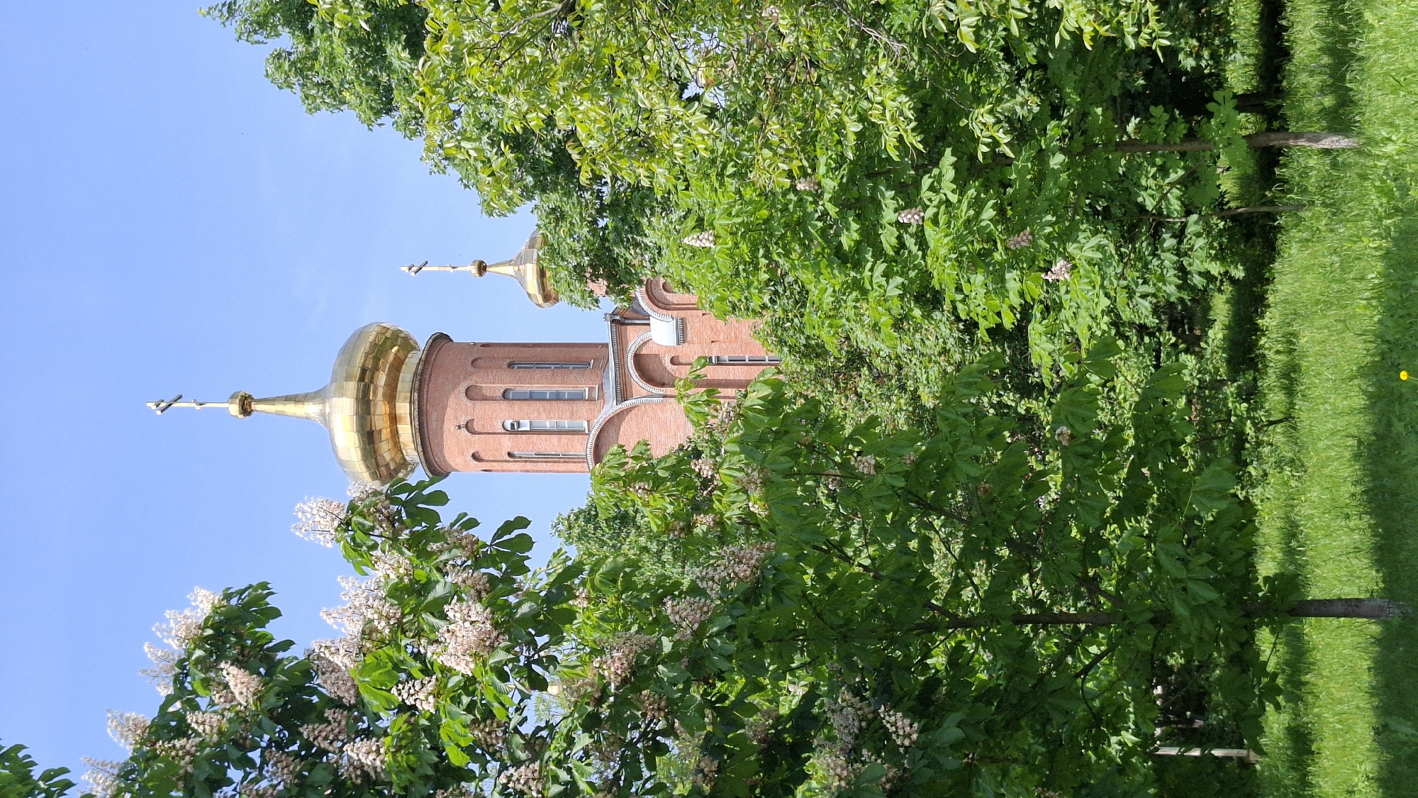 The image shows a brick building with a golden dome and cross. The building is partly obscured by trees and vegetation. There is blue sky visible in the background. It is an outdoor scene on a sunny day.