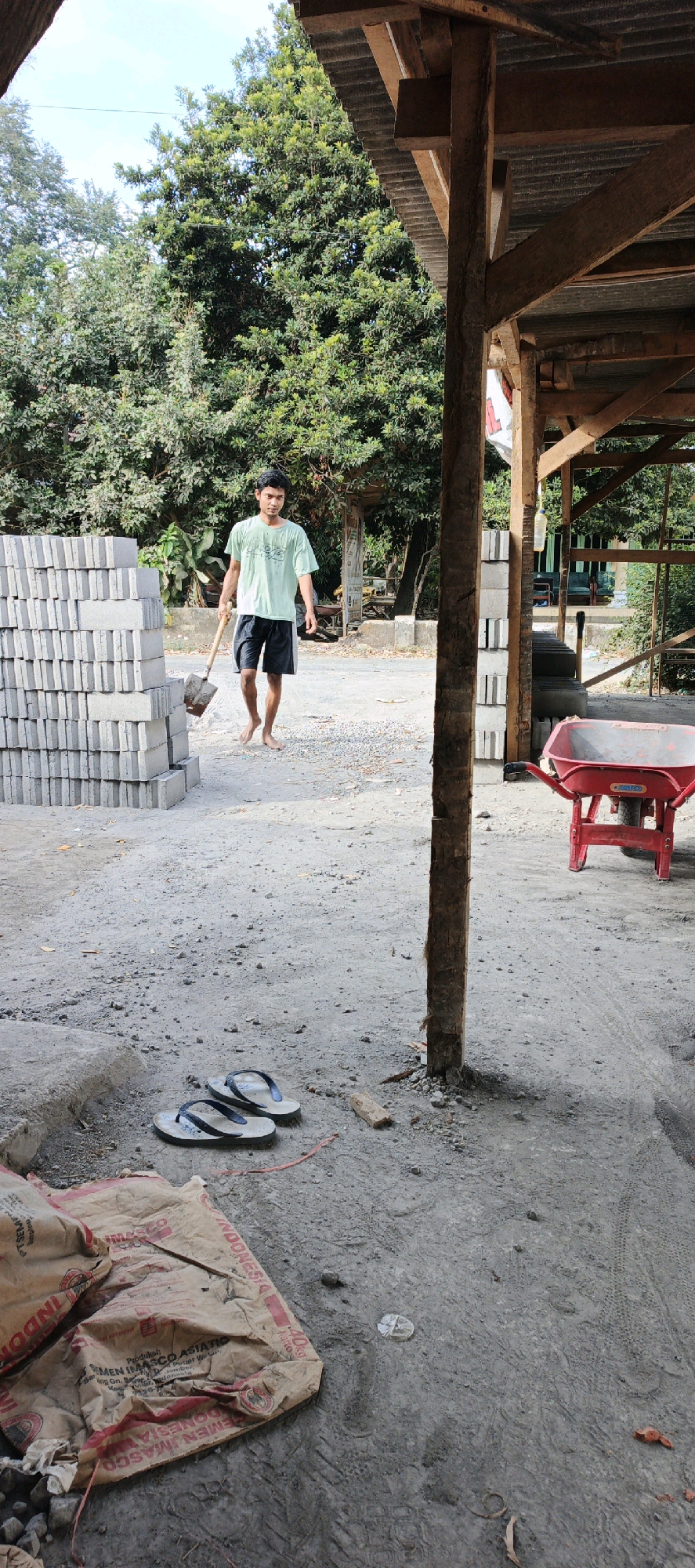A young man, barefoot, walks across a dusty, outdoor construction site in Kasiyan, Indonesia, carrying a shovel. He wears a light green t-shirt and dark shorts. To his left, a tall stack of gray concrete blocks stands neatly piled. In the foreground, a pair of flip-flops rests on the ground near a large, brown paper bag with red text, likely containing cement. Further back, a red wheelbarrow is visible. The scene is partially covered by a wooden-framed structure with a corrugated metal roof. Lush green trees form a backdrop against a bright sky, suggesting it is daytime.