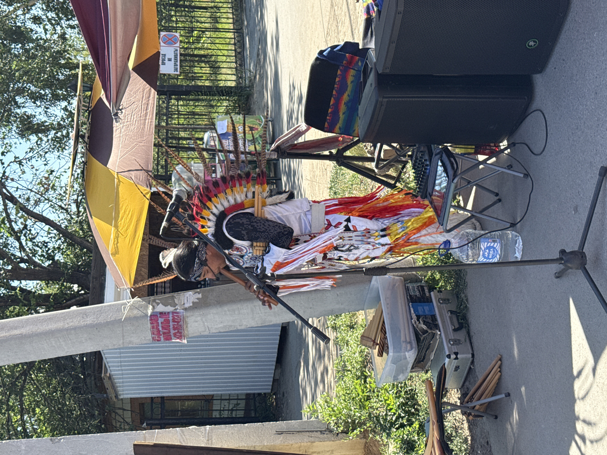 The image depicts an outdoor scene in Yarovoye, Russia, likely during daylight hours. A person dressed in traditional Native American attire, complete with a feathered headdress and fringed clothing, is playing a wooden flute. The individual stands near a concrete pole with several signs attached. Surrounding the performer are various objects associated with a performance or street vending, including a folding table with a digital mixer and tablet, two large black speakers, a cooler containing musical instruments (likely pan flutes), and several folded chairs. In the background, a chain-link fence and trees suggest a public space, possibly near a park or market. The overall impression is that of a street performer set up for an outdoor event.