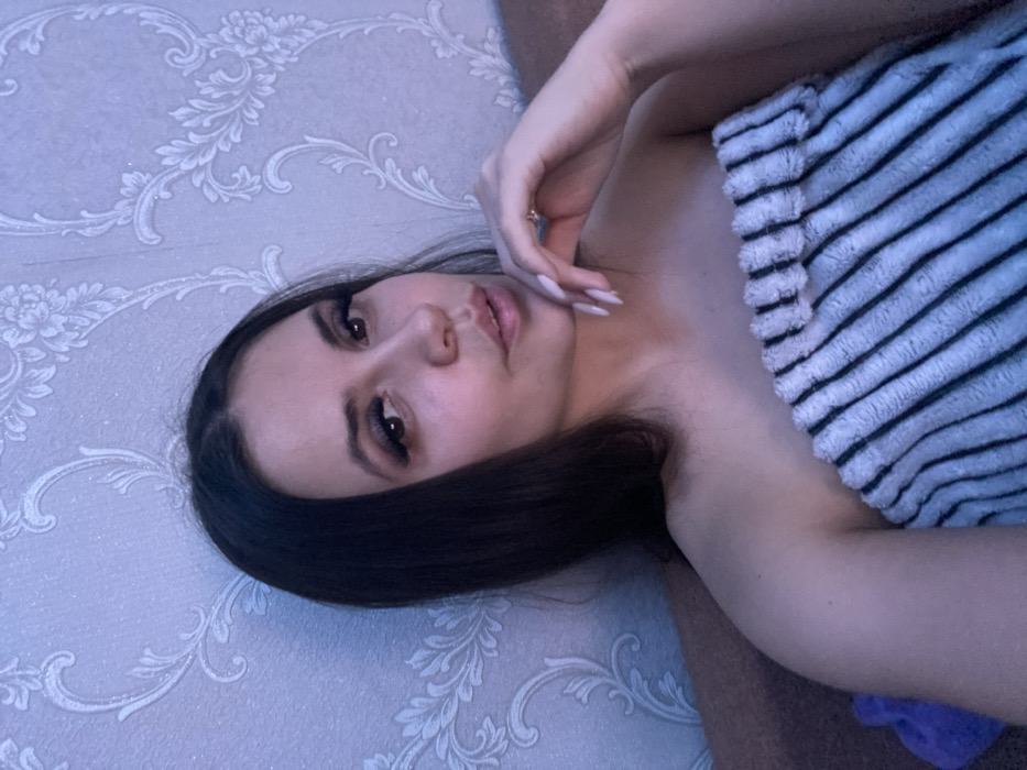 A young woman poses, wrapped in a striped towel. Her long, dark hair falls beside her face, adorned with prominent eyelashes. One hand rests near her face, showcasing long, manicured nails. The background features a patterned wallpaper in light shades. The lighting is soft, contributing to the overall serene and intimate atmosphere of the photograph.