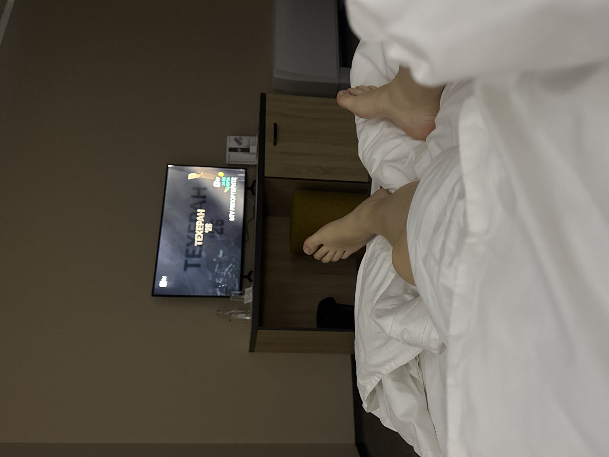The image depicts an interior room, likely a hotel room, in Pleven, Bulgaria, viewed from a supine position in a bed. In the foreground, a person's bare feet are visible, partially covered by white, crumpled bedding. A flat-screen television is centered in the midground, displaying a program from the Bulgarian channel "btv" with Cyrillic text, including "ТЕХЕРАН" (Tehran) and "btv РЕПОРТЕРИТЕ." Below the television, a dark wooden console table holds a clear glass bottle of water, a drinking glass, and a small rectangular object. A dark waste bin and a mustard-yellow cylindrical stool are positioned beneath the table. The room's walls are plain beige, and a white appliance, possibly an air conditioning unit, is partially visible on the far right. The scene is dimly lit, with the television screen providing the primary illumination.