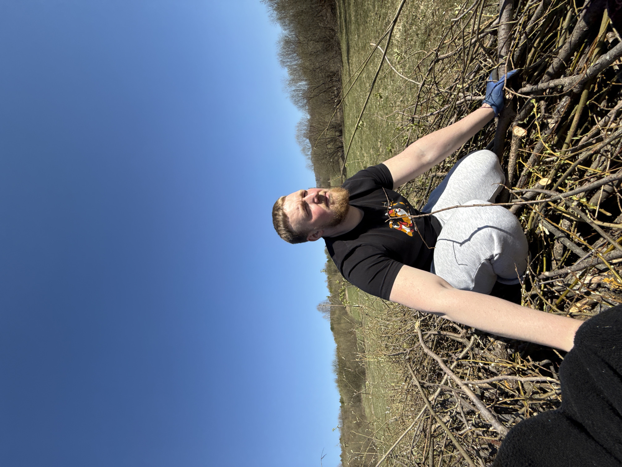 The image shows a man with a beard and wearing a black t-shirt with a graphic on it, light grey sweatpants, and blue gloves. He is kneeling among a pile of branches and sticks, possibly gathering or handling them. The background includes a grassy field and trees, under a clear blue sky. The location is in the city of Nizhnyaya Chermoda, Russia. Another individual, dressed in black, is partially visible in the lower right corner of the image.