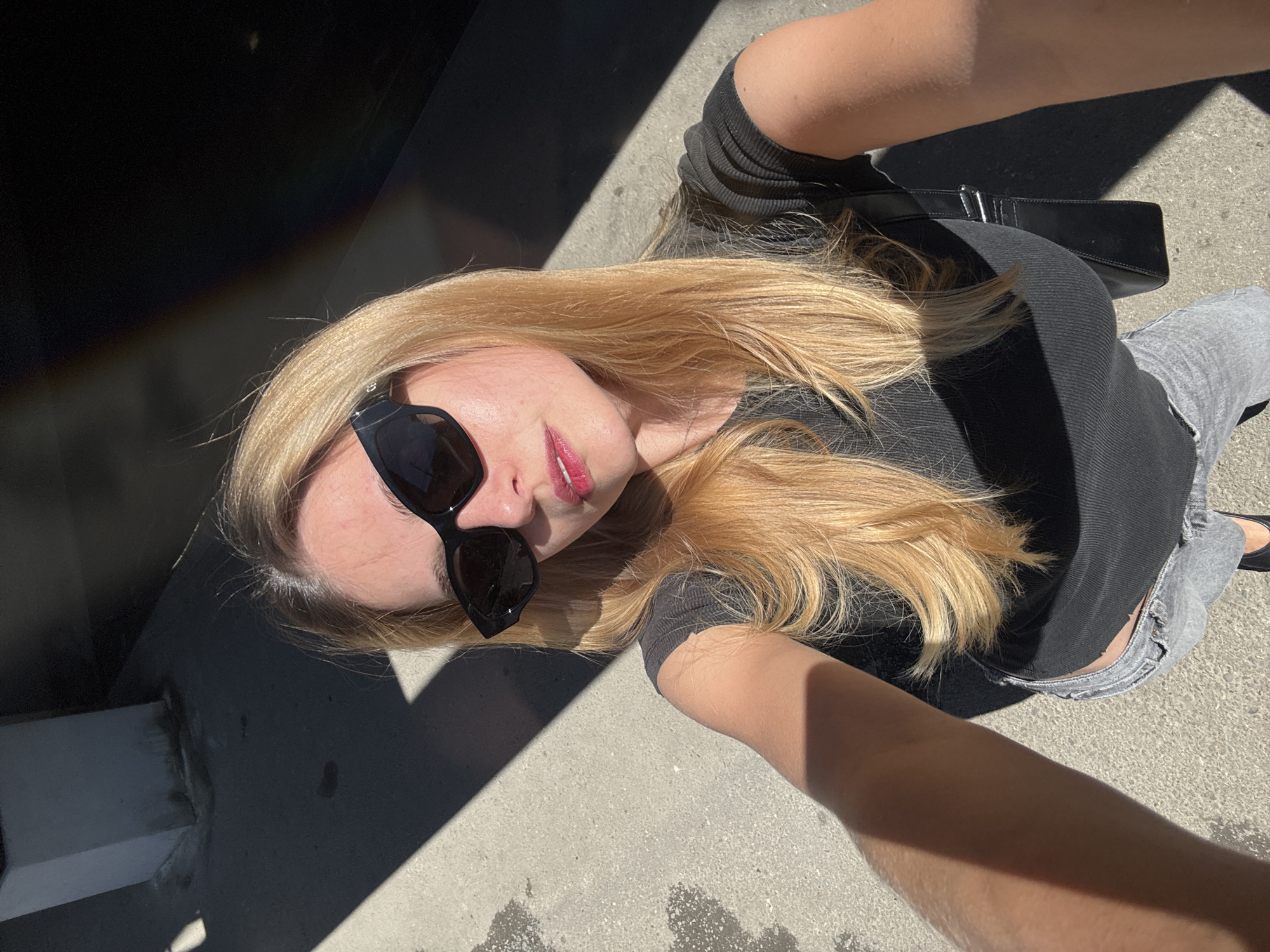 A blonde-haired woman wearing sunglasses, a black top, and gray jeans is pictured in an overhead shot. She is holding the camera and appears to be taking a selfie. She is standing on a concrete surface. Parts of a concrete structure are visible in the left background. The lighting creates strong shadows. The location is Unknown, Unknown.