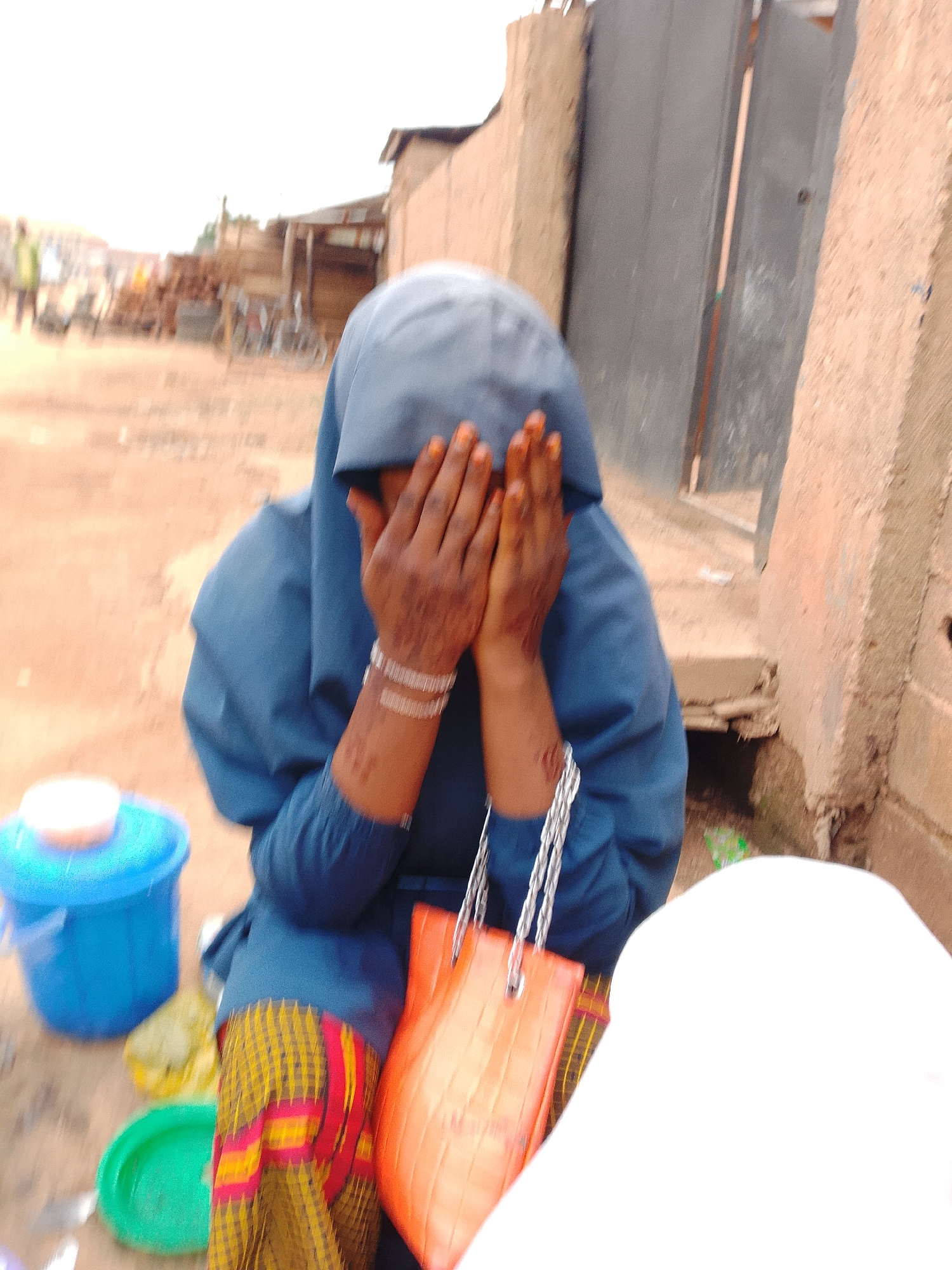 A person wearing a blue head covering and blue robe, with a patterned skirt of yellow, red, and black, sits on the ground. Their hands are covering their face, and they are wearing bracelets on their wrists. An orange purse with silver straps hangs from their arm. To the left of the person is a blue bucket and a green dish, with a yellow plastic bag on the ground. In the background, there are structures that appear to be market stalls, with wooden beams and corrugated metal roofing. The ground is a sandy, reddish-brown color. The scene is outdoors, in what appears to be a marketplace or a village street.