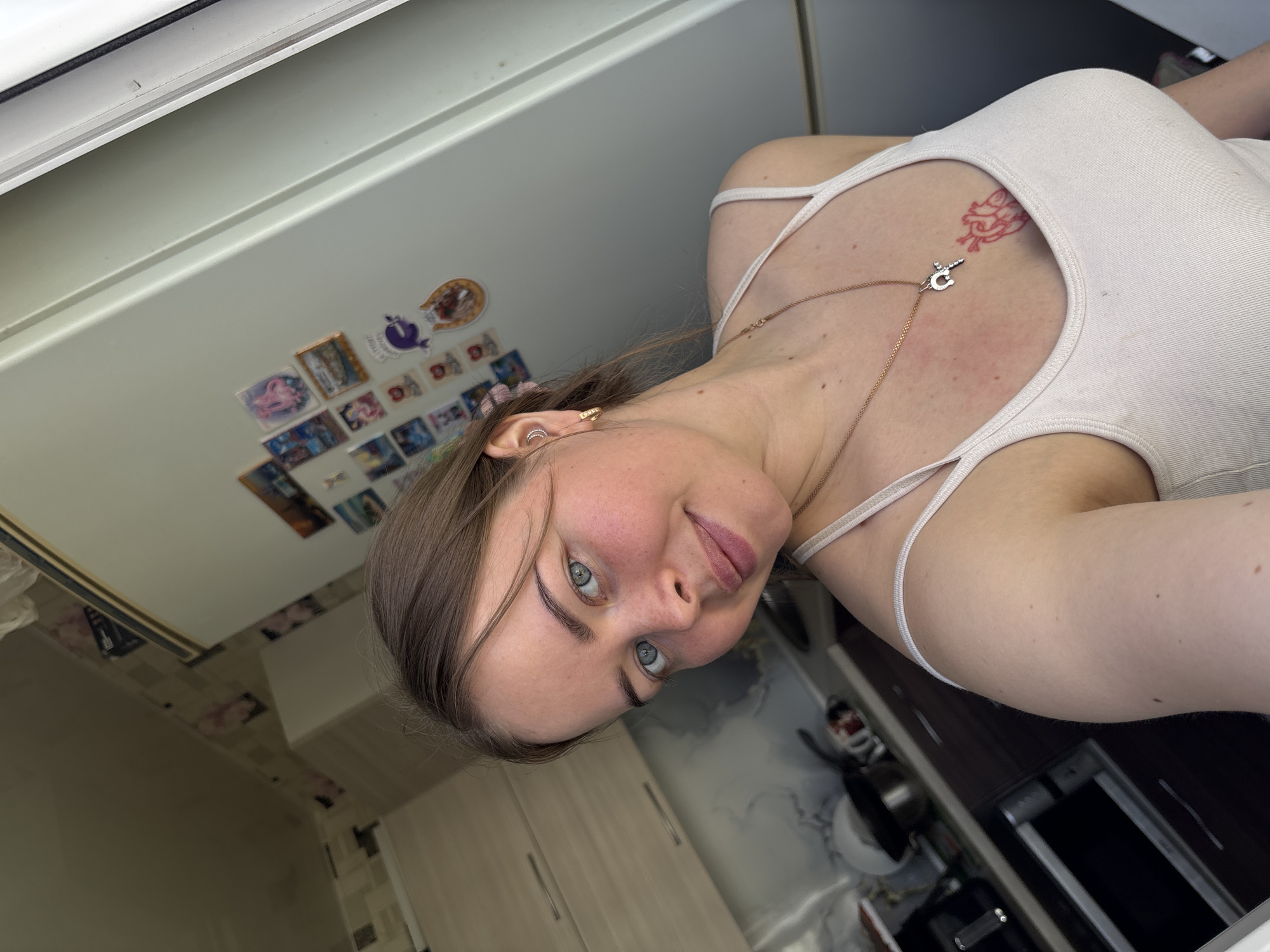 A woman with fair skin, light brown hair, and blue eyes is positioned in the center of the frame, wearing a white tank top and a gold necklace with a pendant. A red tattoo is visible on her upper chest. She looks directly at the camera with a slight smile. The background suggests an interior space, possibly a kitchen. The upper left area has a window frame and a wall covered with small images or stickers. Below the woman, the background includes kitchen cabinets, tiles, and a sink area. The angle of the shot is slightly tilted. While the image contains clues regarding the location (potentially an apartment kitchen), there is no information that confirms the location as Izhevsk, Russia.