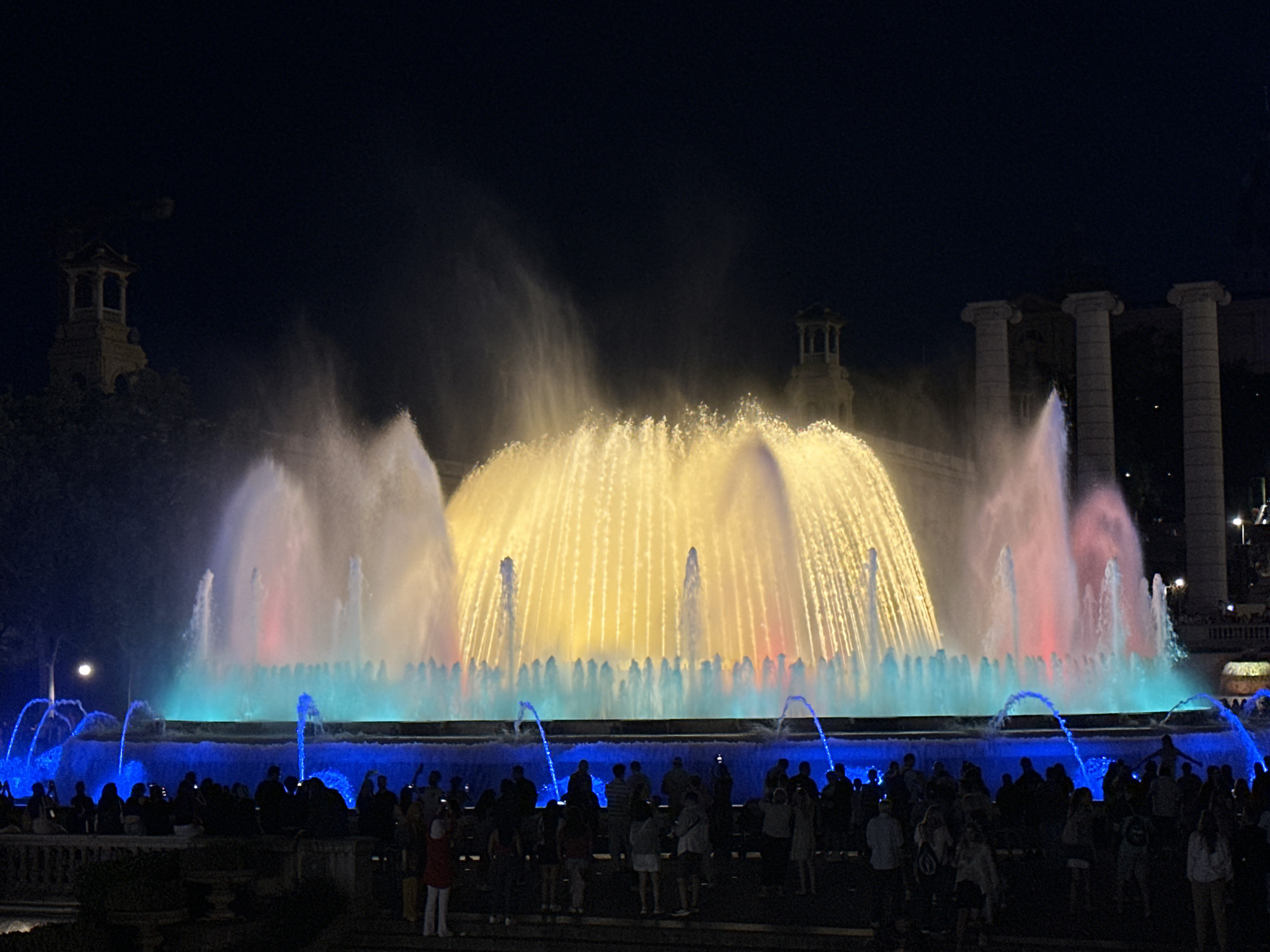 This detailed image captures the Magic Fountain of Montjuïc in Barcelona, Spain, at night, during a synchronized light and water show. The multi-tiered fountain is the central subject, displaying vigorously illuminated water jets. The primary illuminated water columns in the center are bright yellow and gold, flanked by jets of red, pink, and light blue/turquoise. The lowest tier of the fountain features deep blue illuminated water and numerous smaller blue jets arching upwards.
In the foreground, a large crowd of people, predominantly silhouetted against the bright fountain lights, occupies multiple stepped terraces. Many individuals are standing, observing the spectacle, and some are holding up mobile devices, likely recording or photographing the event.
The background reveals architectural elements characteristic of the Montjuïc area. To the far left, a distinct, multi-level tower structure with arched openings is partially visible against the dark sky. To the right, several large, classical columns, consistent with the facade of the Palau Nacional or the flanking structures leading to it, stand prominently. The sky is dark, indicating nighttime conditions, with no visible clouds. The overall scene depicts a vibrant public gathering enjoying an illuminated fountain display.