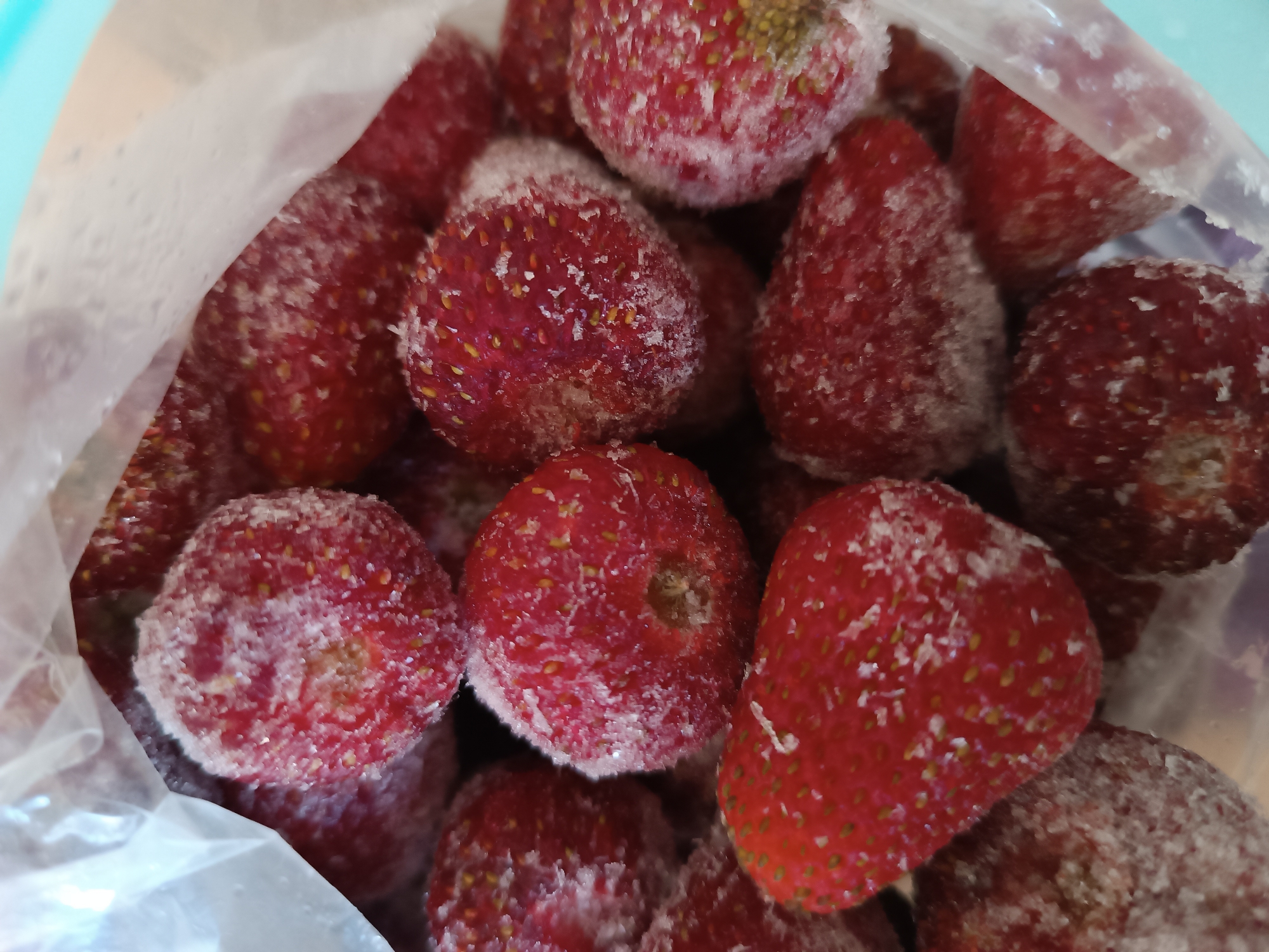 A close-up, overhead shot shows a pile of frozen strawberries inside a clear plastic bag. The strawberries are bright red and covered in a layer of frost, giving them a white, powdery appearance. The bag is slightly translucent and appears to be made of thin plastic. Some of the strawberries are whole, while others appear to be cut in half. The background is a pale, blurry blue. The image is well-lit, with the frost on the strawberries catching the light and creating a slightly glittering effect.