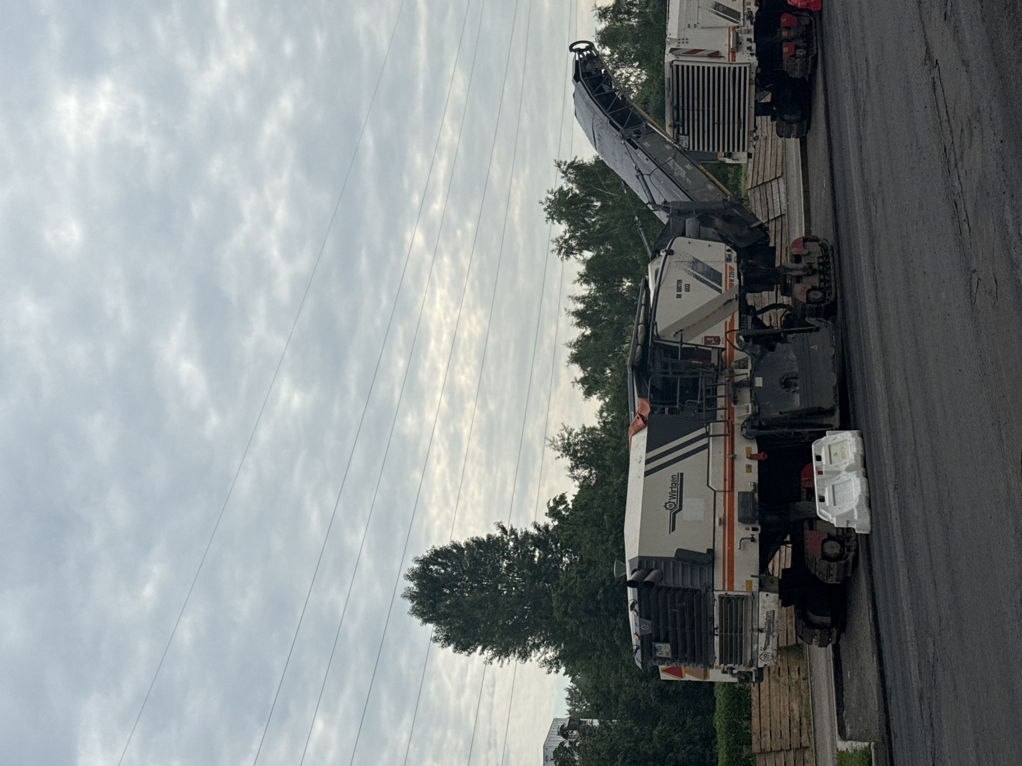 The image depicts a road milling machine, likely a Wirtgen brand, operating on asphalt. The machine is white with black and orange accents and has caterpillar tracks. A conveyor belt extends from the machine, indicating it is actively removing material from the road surface. Two white plastic barriers are placed on the asphalt near the machine. In the background, a line of trees separates the road from a building and power lines are visible against an overcast sky. The scene suggests ongoing road construction or maintenance work. While the image does not explicitly show the city of Moscow, Russia, road milling machines of this type are commonly used in urban infrastructure projects.