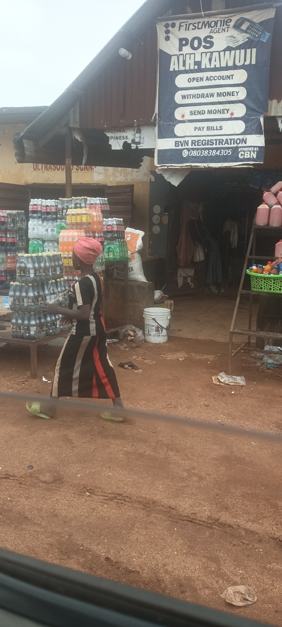 The image shows a street scene in Kassa Market, Nigeria. In the foreground is an unpaved dirt road. In the midground, a woman wearing a striped garment and a head covering is walking. She wears lime green sandals. To her left are stacks of bottled drinks. To the right, behind the woman, is a shop with a sign that reads "Firstmonie Agent POS ALH. KAWUJI," advertising services such as opening accounts, withdrawing and sending money, paying bills, and BVN registration, approved by CBN, with a phone number. A white bucket and a bag labeled "FISH STARTER PELLETS" are nearby. Inside the shop, hanging clothing can be seen. A metal structure with items, possibly food or household goods, in a green basket is also visible. The sky appears overcast.