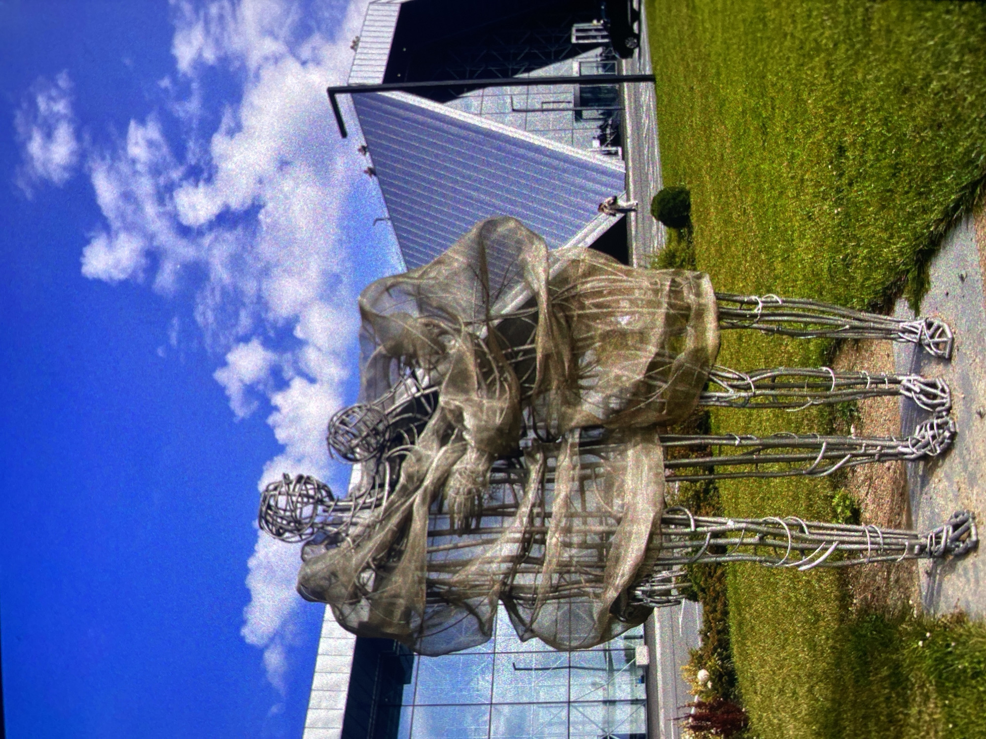 The image shows a metal sculpture of several intertwined figures located in Saint Petersburg, Russia. The sculpture is positioned on a concrete path beside a grassy area. A modern building with a glass facade and a sloping roof is in the background. The sky is blue with scattered white clouds. There are people visible in the distance near the building.