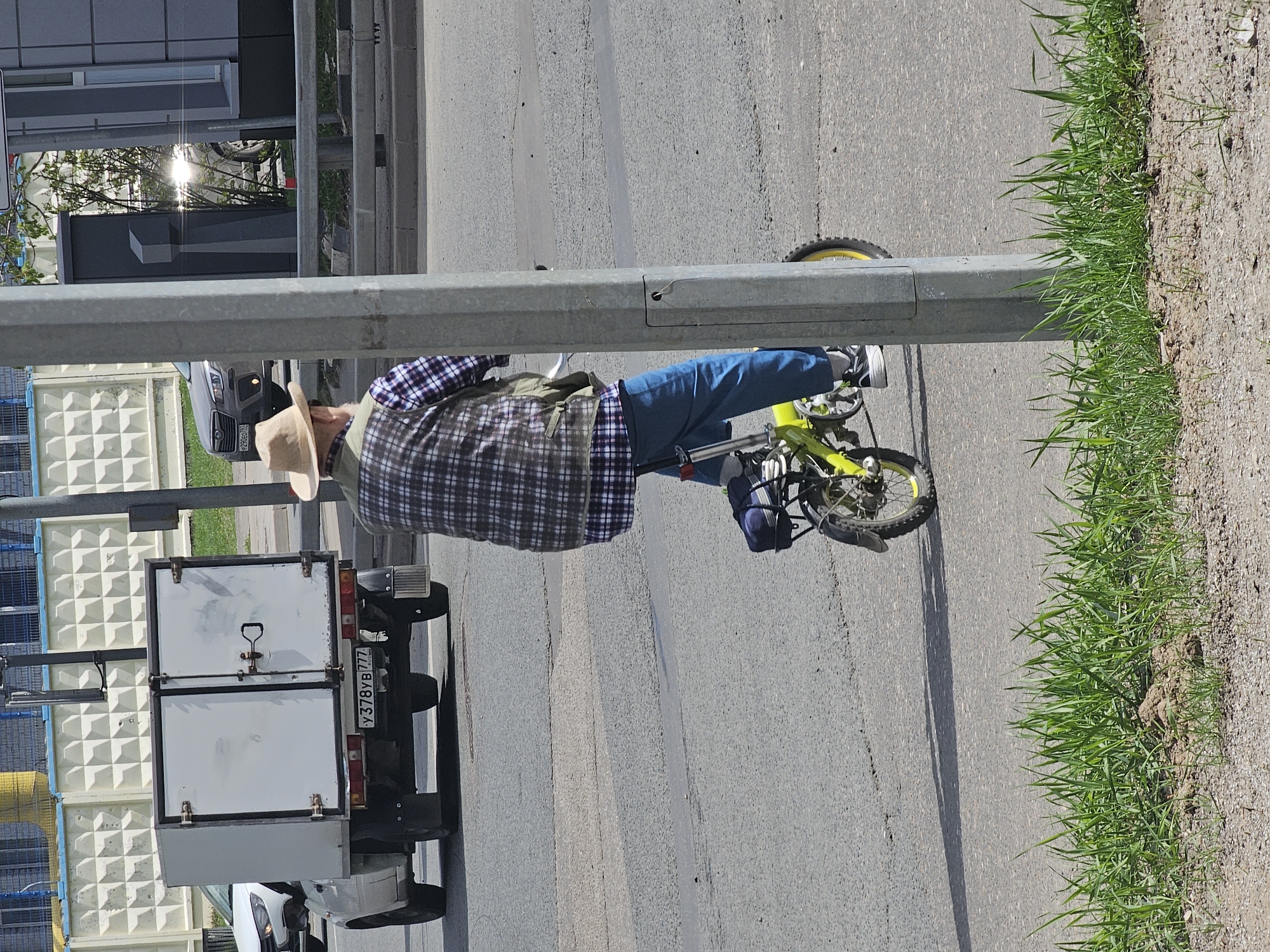 A person wearing a straw hat, plaid shirt, and jeans is riding a yellow bicycle on a paved road. A white truck with a boxy container attached to the back is to the left of the person. The license plate of the truck reads "Y378YB777." A metal pole is in the foreground. Grass and dirt are to the right of the road. The location is Khimki, Russia.