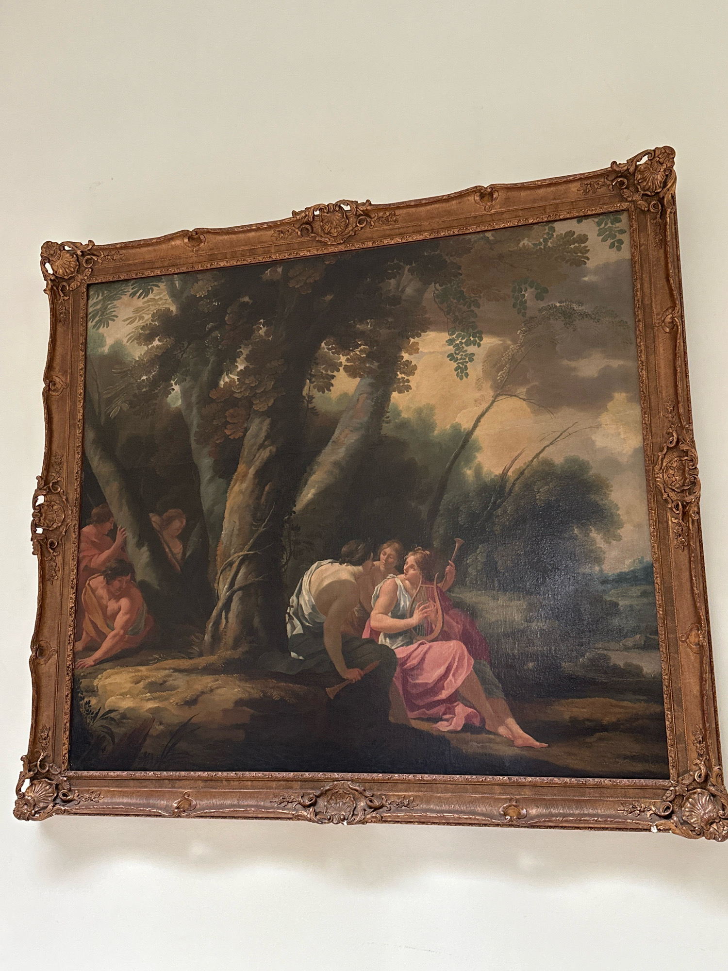 This is a full-shot, eye-level photograph of a large, ornately framed oil painting hanging on a plain white wall. The painting depicts a pastoral scene with several figures. In the foreground, on the right, a woman with light skin and dark hair is seated and playing a lyre. She is wearing a pink draped garment. To her left, leaning towards her, is a man with light skin and dark hair, wearing a light blue garment. His left arm is around the woman. Behind them, to the left, are several other figures partially obscured by trees. One man in the lower left is crouching, wearing a tan garment. Further back and to the right, there are more trees and foliage, with a landscape featuring water and distant hills under a cloudy sky in the background. The painting is displayed in a gilded, decorative frame with scrolling motifs and shell-like embellishments at the corners. The photograph is taken from a slightly angled perspective, with the top of the frame reaching higher than the bottom. There is no information in the image to suggest it is located in Belgrade, Serbia.