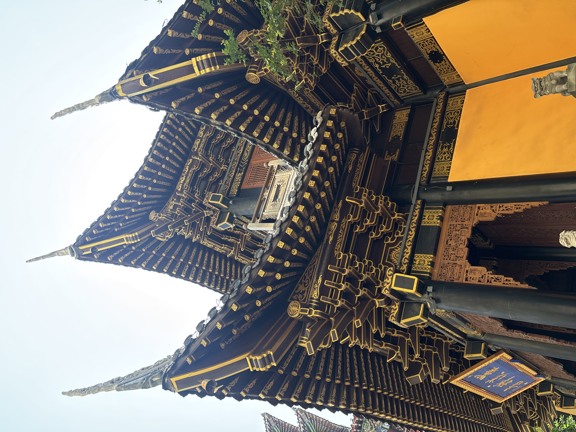 The image shows a close-up, upward-angled view of the ornate roof of a traditional Chinese temple located in Yuzhong District, China. The roof is constructed of dark wood with intricate golden decorative patterns and features multiple tiers with upturned eaves, characteristic of ancient Chinese architecture. Several golden spires rise from the corners of the roof, some adorned with what appear to be decorative figures or carvings. Small bells hang from some of the eaves. The exterior walls of the temple building visible in the lower part of the image are painted a bright orange-yellow. A blue banner with golden Chinese characters hangs from the left side, and a small stone lion statue is visible in the lower right corner. Green foliage from a nearby tree can be seen to the right. The sky above is bright and overcast. There are no people or animals actively engaged in any discernible actions within the frame.