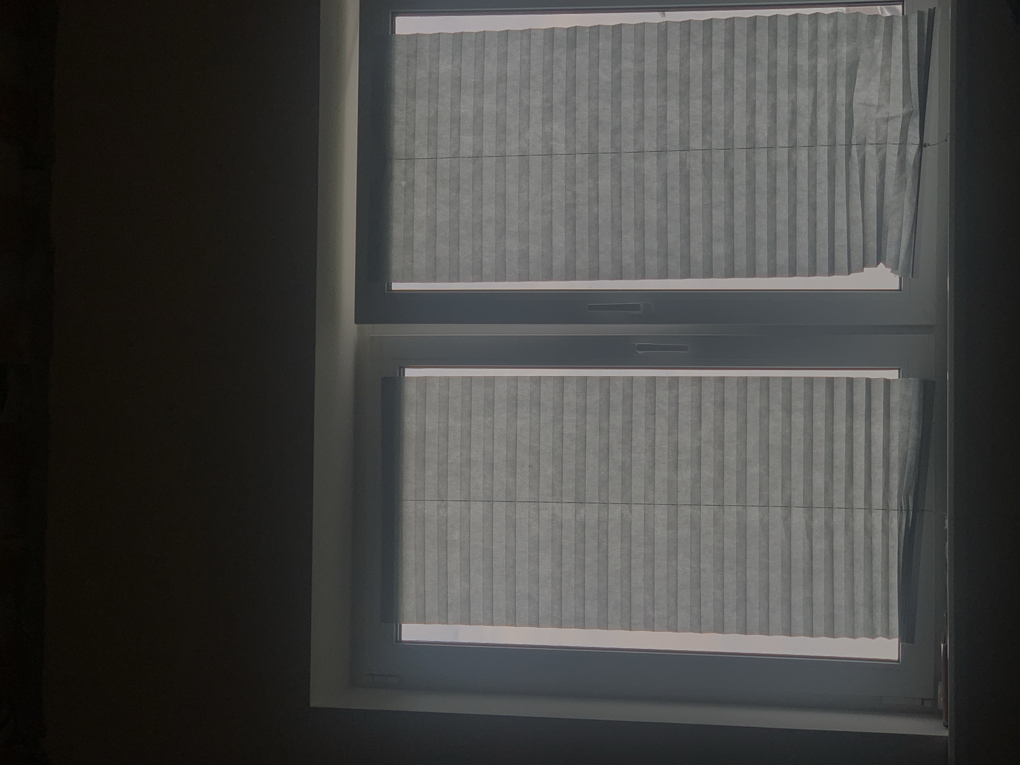 A dimly lit interior shot shows a closed, white-framed window with two pleated blinds. The blinds are a muted, grayish-blue color and are partially covering the window panes, allowing some diffused light to filter through. The left blind appears to be fully closed, while the right blind is slightly open at the bottom, revealing a hint of the bright, possibly outdoor, light beyond. The window is divided vertically by a white mullion. The wall above the window is dark and appears unfinished. The windowsill is dark and has a slight sheen. The overall atmosphere is somber and quiet. This scene could be located in an apartment or residential building in Minsk, Belarus, given the typical window and blind styles common in the region.