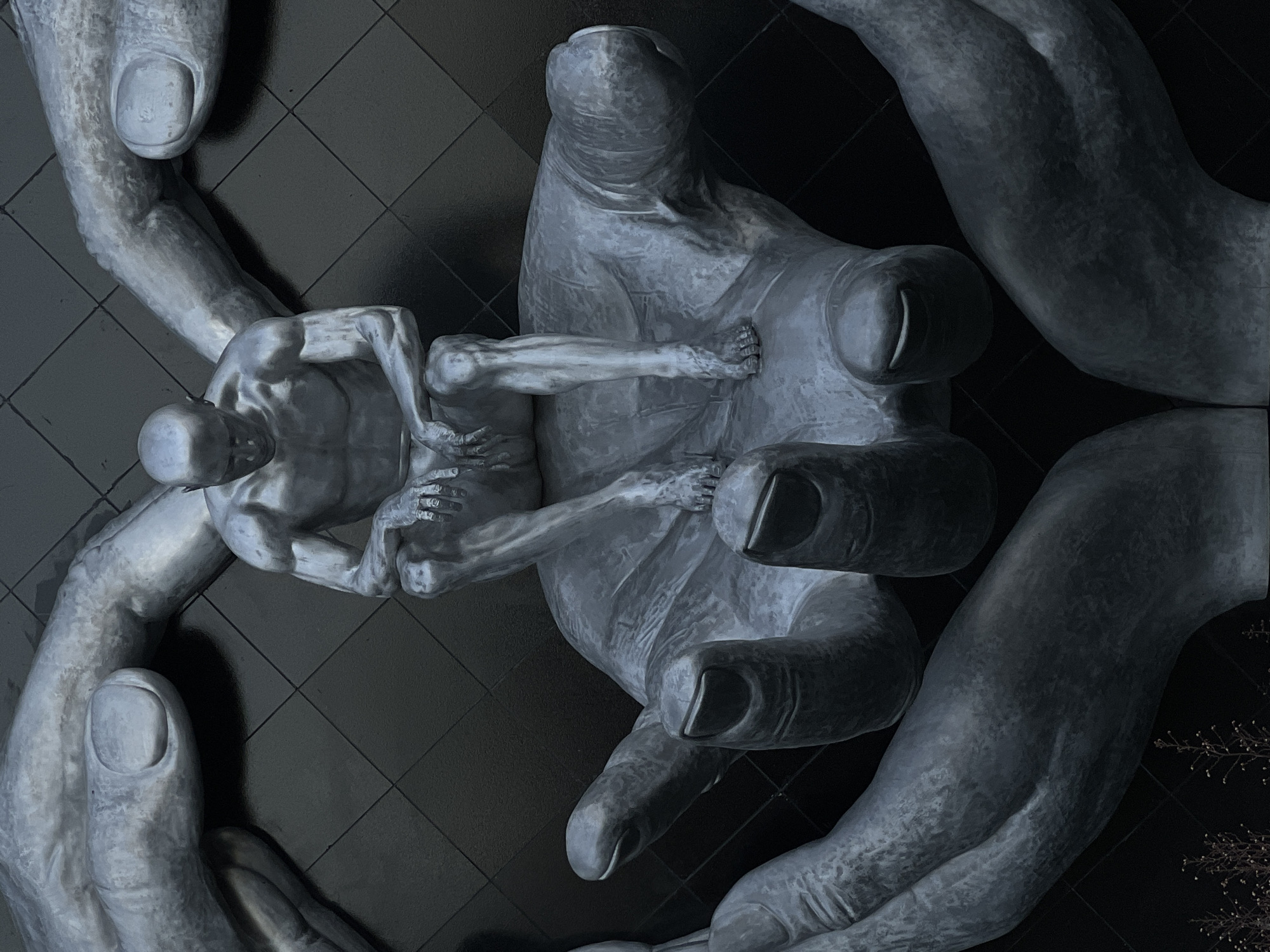 The image shows a large, metallic sculpture located in Krasnodar, Russia. The sculpture depicts a figure of a man sitting in a contemplative pose, supported by several large, cupped hands. The man is bald and appears to be made of the same metallic material as the hands. His arms are resting on his knees, and his head is bowed. The hands are positioned around him, appearing to cradle or protect him. The background is a dark, tiled surface. The sculpture is monumental in scale, and the metallic finish gives it a somber and imposing appearance. The overall impression is one of vulnerability and support, with the large hands acting as a symbol of protection or care for the solitary figure.