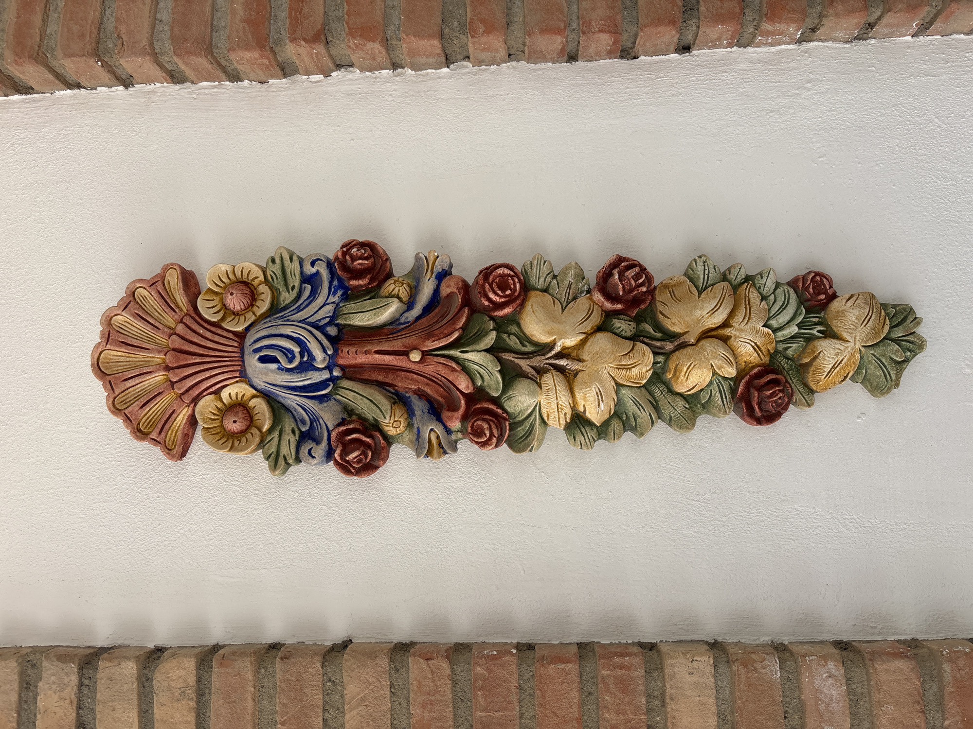 The image shows a colorful, ornate stucco relief decoration mounted on a white wall. The relief depicts a fan-shaped element at the top, with flowing ribbons and floral motifs, including roses and what appear to be stylized leaves or petals, extending downwards. The colors are vibrant, featuring shades of red, yellow, blue, green, and pink. The decoration is flanked by two brick pillars, one on the left and one on the right, suggesting it might be an architectural detail on the exterior of a building. The bricks are of a reddish-brown hue and laid in a stretcher bond pattern. The white wall has a slightly textured surface. This type of decorative relief is commonly found in the historic architecture of Orihuela, Spain, particularly on buildings in the city center or in the older neighborhoods. Without specific landmarks, pinpointing the exact location within Orihuela is not possible from this image alone.