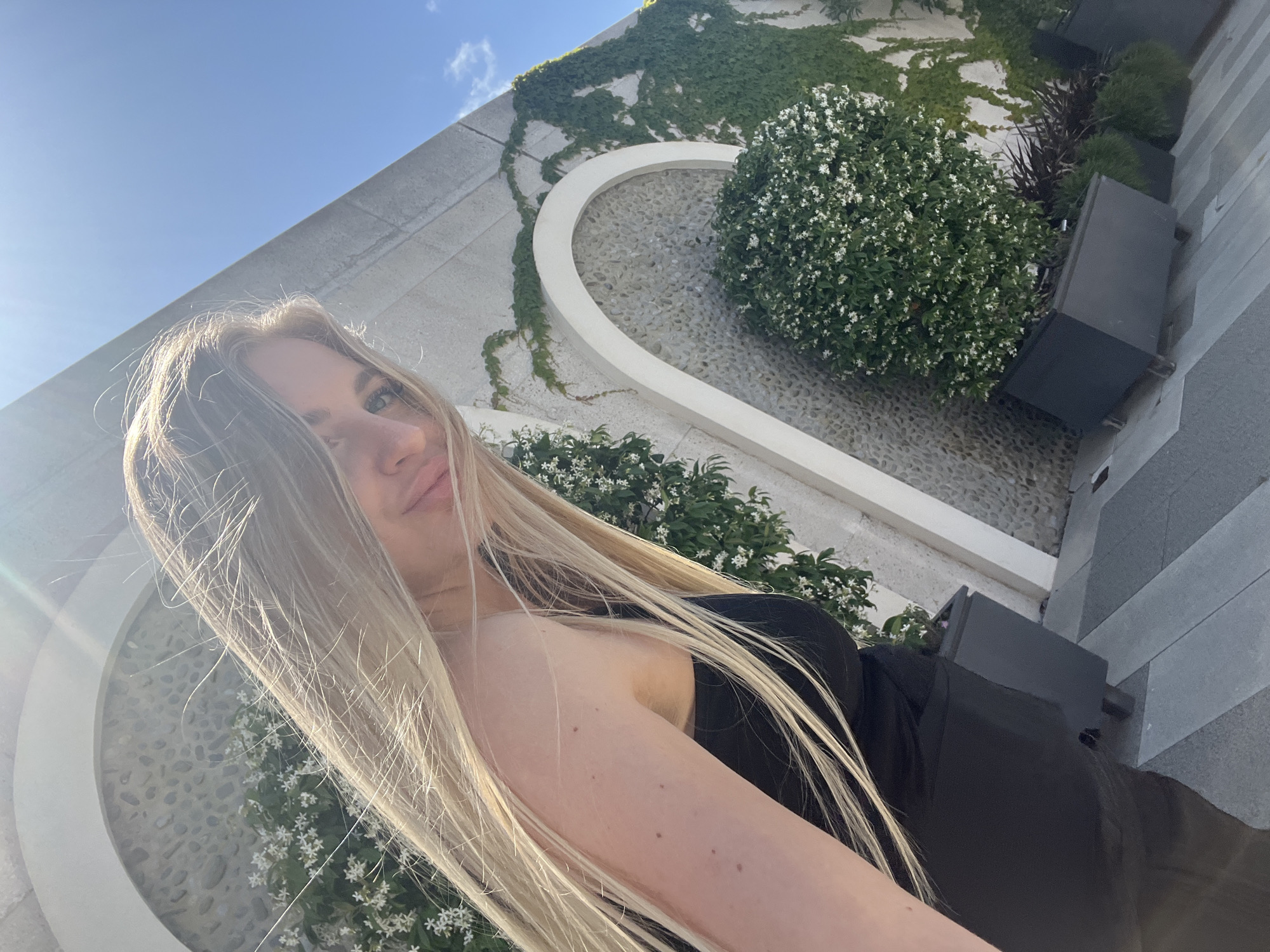 A woman with long blonde hair is in the foreground of the image. She is wearing a black top. Behind her is a modern architectural structure with integrated greenery. A wall covered in ivy is visible, as well as cultivated bushes with small white flowers. There are also dark gray planters with various plants. The surface of the courtyard or garden is pebbled and paved with gray stone tiles. The sky is blue with some light clouds. The setting appears to be an upscale outdoor space in Radovići, Montenegro, as indicated by the architectural style and landscaping.