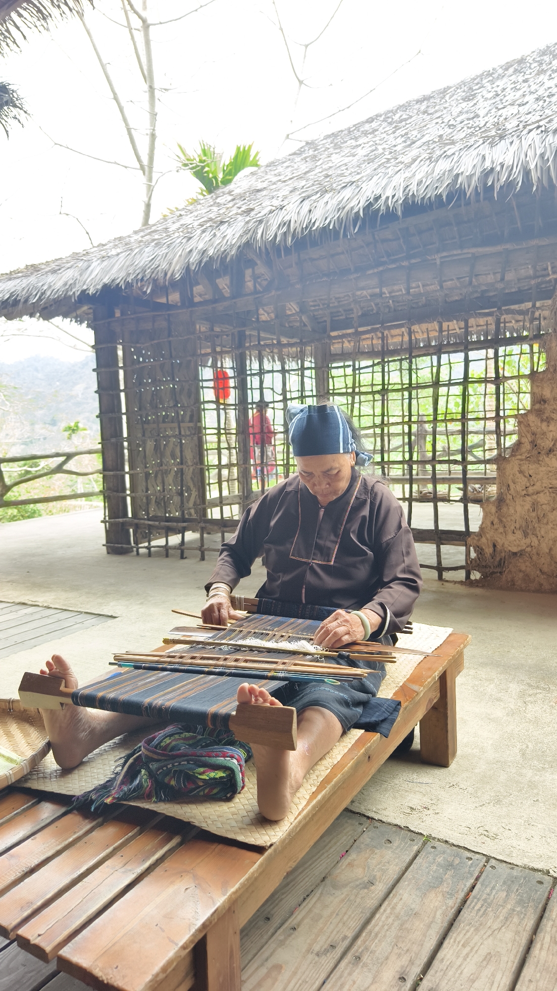 An elderly woman, identified by traditional attire including a dark headscarf, dark long-sleeved shirt with light trim, and dark skirt, is depicted seated on a low wooden bench, engaged in weaving on a traditional loom. She wears two light-colored bangles on her left wrist and is barefoot, using her feet to brace part of the loom's tensioning mechanism. The loom features dark blue/black and white threads. A folded, multicolored woven fabric is placed on the bench between her feet.
The scene takes place under a shelter with a prominent thatched roof and rough wooden support poles. The background features a wall constructed from woven bamboo or thin wooden slats, with an exposed red lantern hanging within it. To the right, a section of an earthen or mud wall is visible. A concrete or paved floor is directly beneath the weaving area, transitioning to wooden planking in the immediate foreground. In the distant background, outside the structure, rolling hills or mountains with sparse vegetation are visible under an overcast sky. A person in a red garment is partially visible within the bamboo-walled section of the structure. The location is in Jiyang District, China.