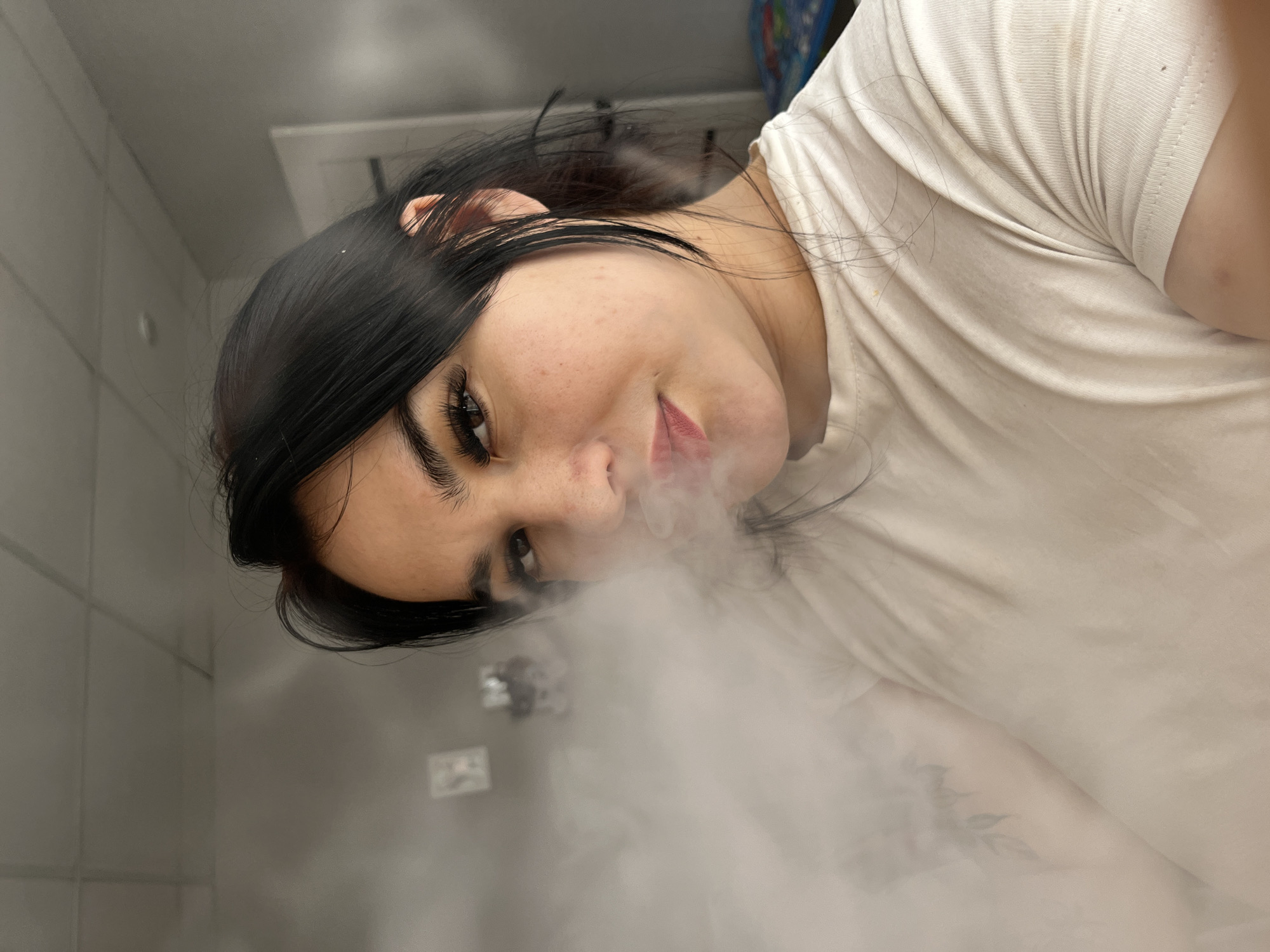 A woman with dark hair and fair skin, wearing a white t-shirt, is partially obscured by a cloud of white vapor. She is looking directly at the camera with a slight smile. The background is a neutral gray, with tiled walls visible on the left and indistinct wall fixtures in the distance. The location could not be determined to be Chaikovsky, Russia from visual information.