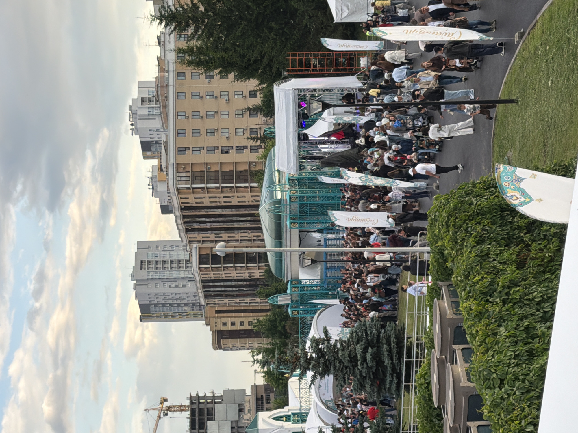This is an outdoor photograph taken in the city of Unknown, Unknown. In the foreground, lush green bushes and manicured grass occupy the bottom portion of the image. Behind the greenery, a large crowd of people is gathered in an open area. Several white tents and a stage structure are visible, suggesting an event or festival is taking place. A teal-colored decorative fence or archway is prominent in the middle ground, adding an architectural element to the scene. In the background, a series of modern, multi-story buildings rise against a cloudy sky. A construction crane is also visible on the left side, indicating ongoing development in the area. The overall atmosphere appears to be lively, with many individuals milling about and interacting.
