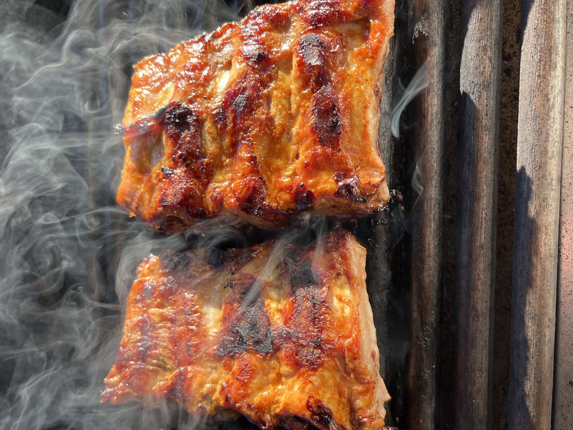 The image depicts two sections of cooked ribs on a grill, emitting smoke. The ribs are browned and appear to have some charring. The grill is made of dark, metallic rods. The setting is not identifiable as Pchelnik, Bulgaria, based on visual information. No people are visible.