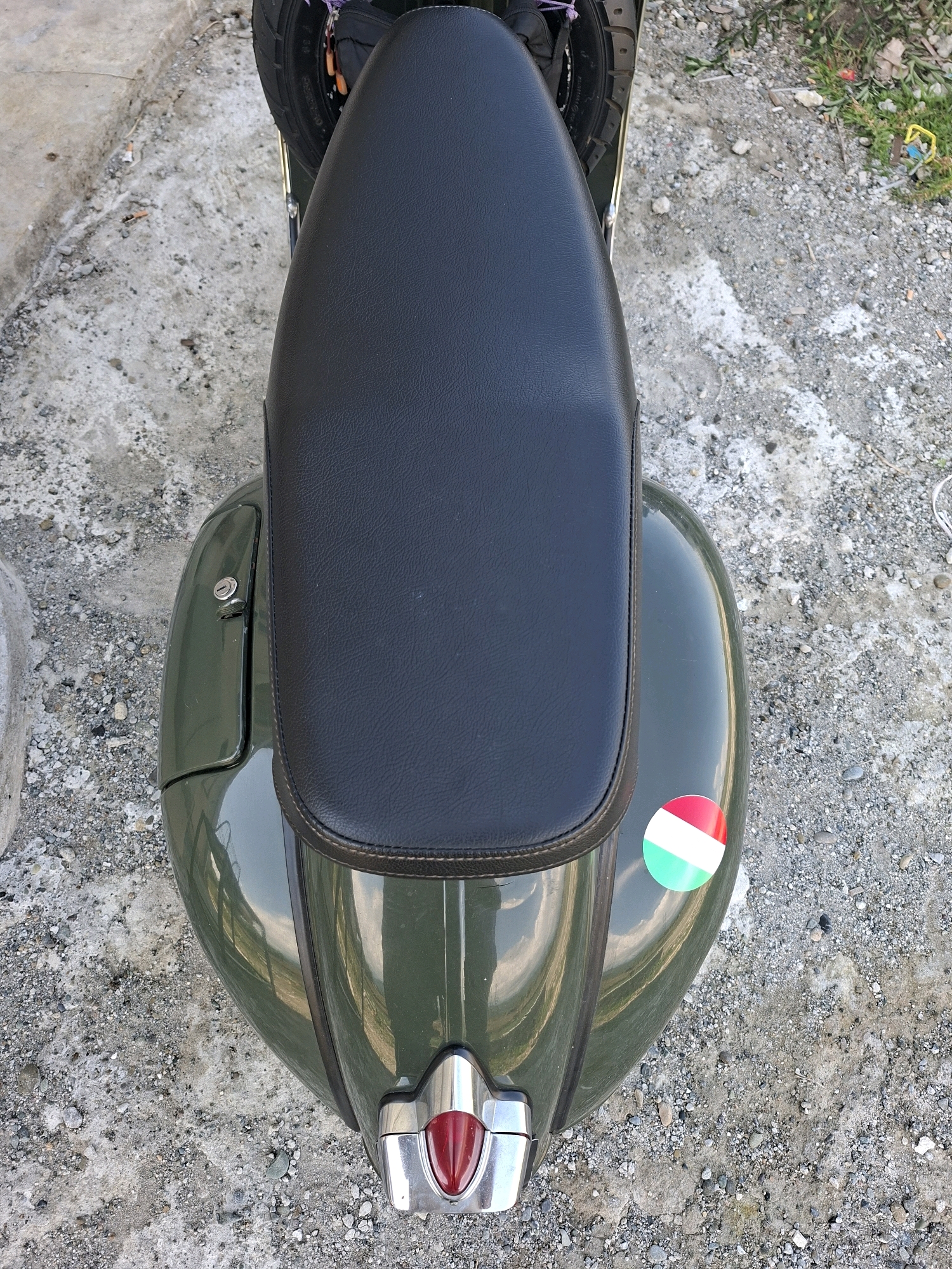 A high-angle, full shot captures the top portion of a dark green vintage scooter parked outdoors on a gravel and concrete surface. The scooter's black seat dominates the center of the frame. To the left, a portion of the rear tire is visible. On the right side of the scooter's body, a circular sticker with red, white, and green stripes, resembling the Italian flag, is affixed. The scooter's rear brake light, a red jewel-like shape encased in chrome, is visible at the bottom of the image. The background consists of rough, uneven ground with scattered pebbles and some dried vegetation, suggesting an outdoor, possibly urban or semi-rural, environment. The scene does not appear to be in Gronggrong, Indonesia, as the visual elements do not suggest that specific location.