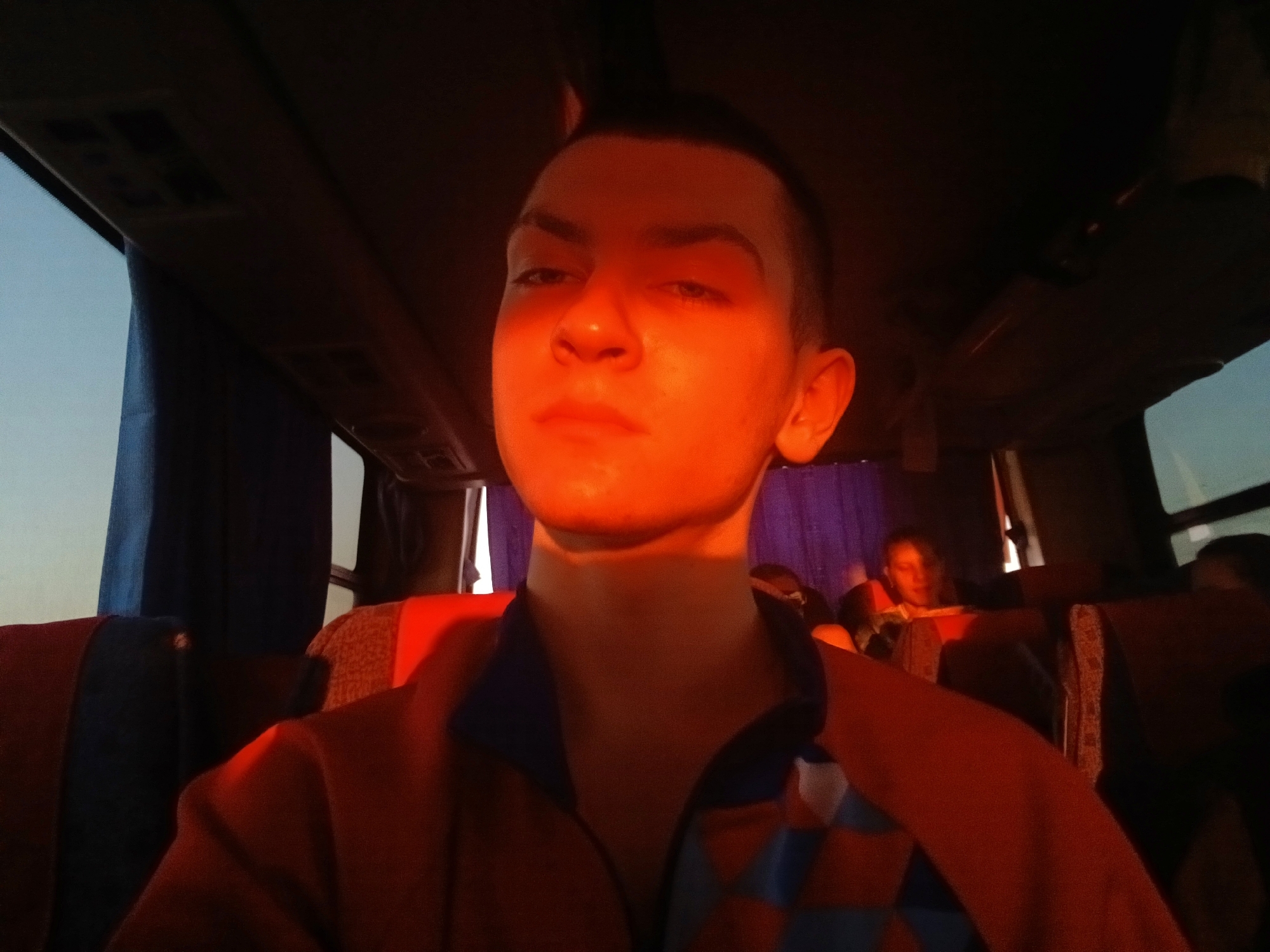 The image shows a young person with short, dark hair sitting on a bus or large vehicle. The subject is facing the camera with a serious expression. The interior of the bus has patterned seats, windows, and overhead storage. The lighting is warm and orange-toned, likely due to the time of day or artificial lighting within the vehicle. Another person can be seen in the background, seated behind the subject. The location of the photo is the inside of a bus in Ustinovo, Russia.