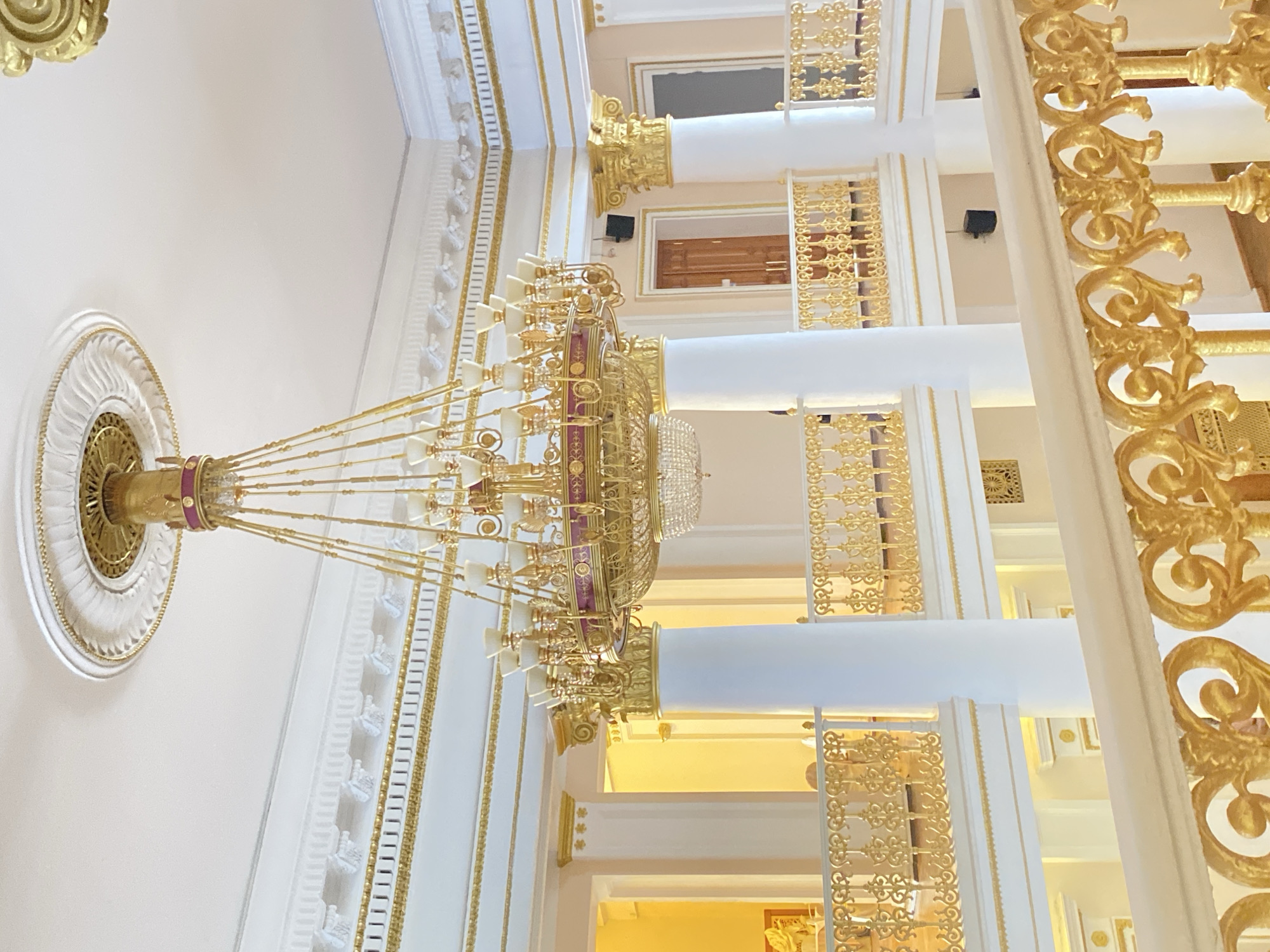 The image captures an opulent interior with a large, ornate chandelier suspended from the ceiling. The ceiling itself is white with intricate decorative moldings. The architecture features white columns with golden accents and filigree, creating a sense of luxury and grandeur. The space appears to be multi-storied, with balconies or upper levels visible through the columns. The overall color palette is dominated by white and gold, further emphasizing the lavishness of the environment. No people are visible in the frame. The location appears to be an upscale building in an unidentified city.