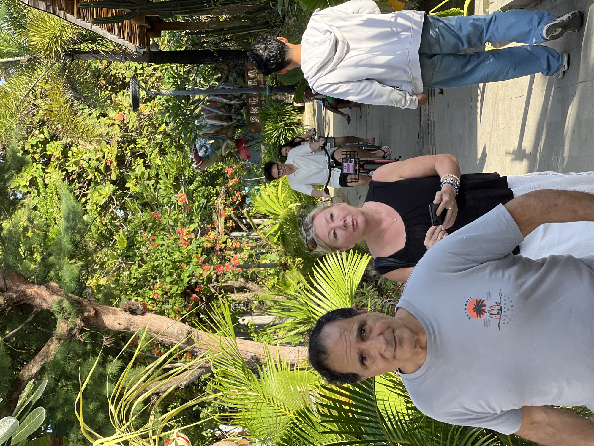 A group of people are walking and cycling on a paved path lined with lush tropical foliage and some structures in Denpasar, Indonesia. In the foreground, a man with dark hair and a grey t-shirt with a "Kuta Bali" graphic is looking at the camera. Beside him, a woman with blonde hair, sunglasses on her head, and a black tank top is holding a smartphone. Further down the path, a young man in a white short-sleeved shirt and black shorts is riding a bicycle. Behind him, another person is also on a bicycle, and a person in a grey hoodie and blue jeans is walking away from the camera. In the background, there are trees, flowering plants, and a building with a wooden roof. A sign that reads "SEGARA" is visible. The environment appears to be a recreational or tourist area, possibly near a beach or resort.