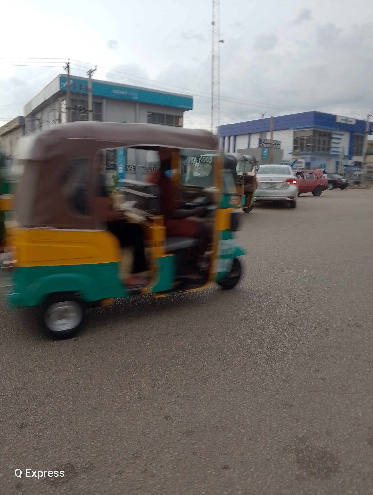 The image captures a street scene in Jos, Nigeria, likely during the day with overcast skies, indicated by the diffused lighting. The foreground is dominated by a moving auto-rickshaw, a common mode of transportation in many parts of the world. The auto-rickshaw is painted in vibrant yellow and green, with a brown canopy. The motion blur suggests it is in transit, and a person, possibly the driver, is visible inside.
Behind the auto-rickshaw, several cars are also on the road, some appearing to be in traffic. Notable among them is a red car with its trunk open. In the background, buildings line the street, suggesting an urban or semi-urban environment. One building has a prominent blue and white facade, possibly a commercial establishment or office. Another building to the left has a lighter colored facade with a blue trim. A tall communication mast is visible in the distance, a common feature in populated areas.
The road surface is asphalt, showing signs of wear and tear. The overall atmosphere is ordinary, depicting a typical day on the road. There is a piece of visible text on the auto-rickshaw that reads "TA-6009". Additionally, in the bottom left corner of the image, the text "Q Express" is present, likely a watermark or branding. The weather appears to be cloudy, contributing to the subdued lighting.