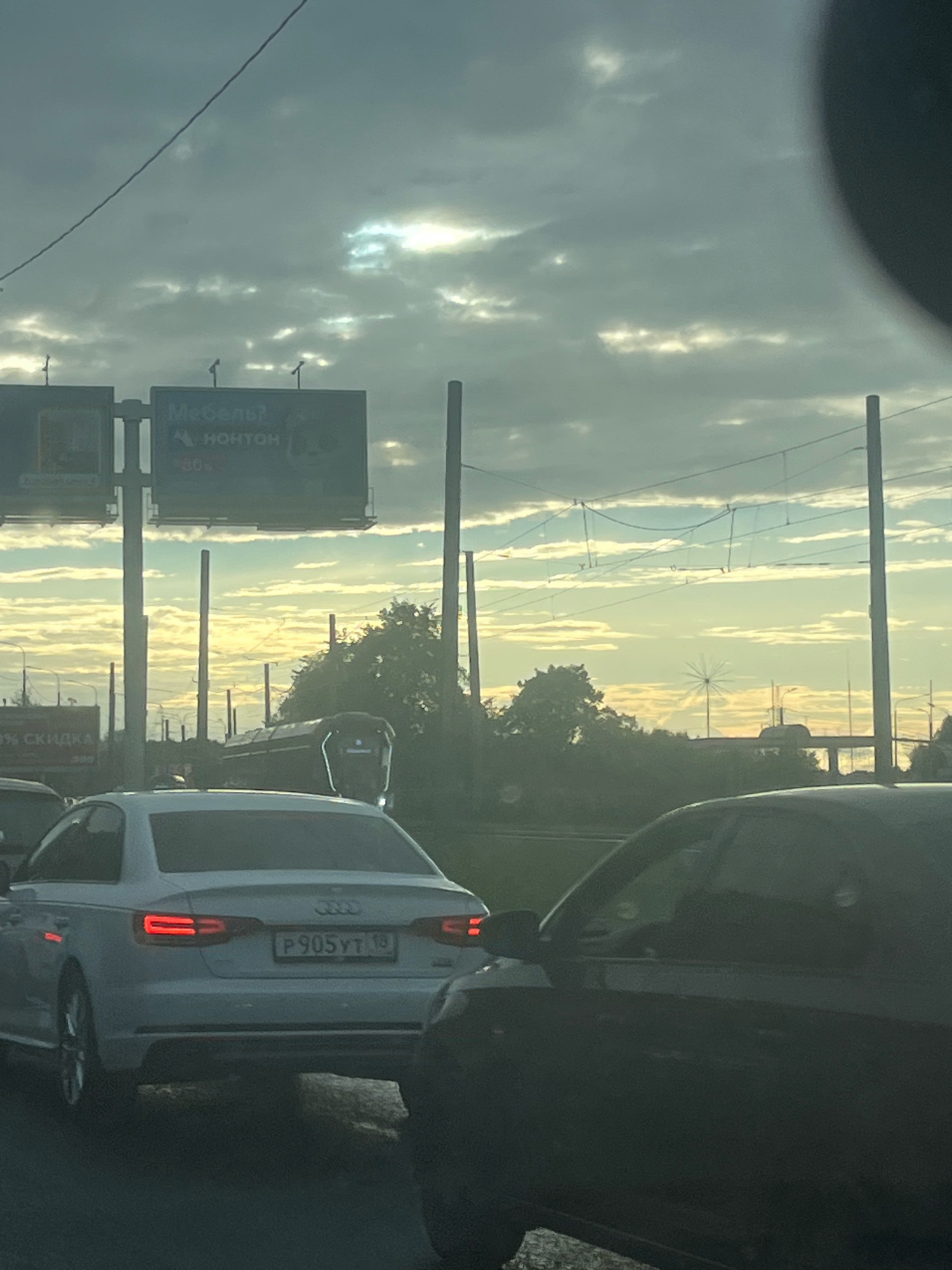 The image captures a view from inside a vehicle, looking forward through the windshield. The scene is a city street with moderate traffic under a cloudy sky with patches of sunlight breaking through. In the foreground, the rear of a white Audi sedan is visible, displaying a Russian license plate that reads "P905YT 19". To the right, the side of a dark-colored vehicle is visible, partially obscuring the view. In the middle ground, there are multiple vehicles in traffic, including a truck with "10% СКИДКА" (10% DISCOUNT) advertised on its side. Billboards are also present, one of which displays "Мебель?" (Furniture?) and "НОНТОН". In the background, trees and utility poles line the street, and a distant structure resembling a Ferris wheel or a large wind turbine is visible against the sky. The overall impression is a typical urban environment during daylight hours, likely in Saint Petersburg, Russia, given the Cyrillic text and license plate format.